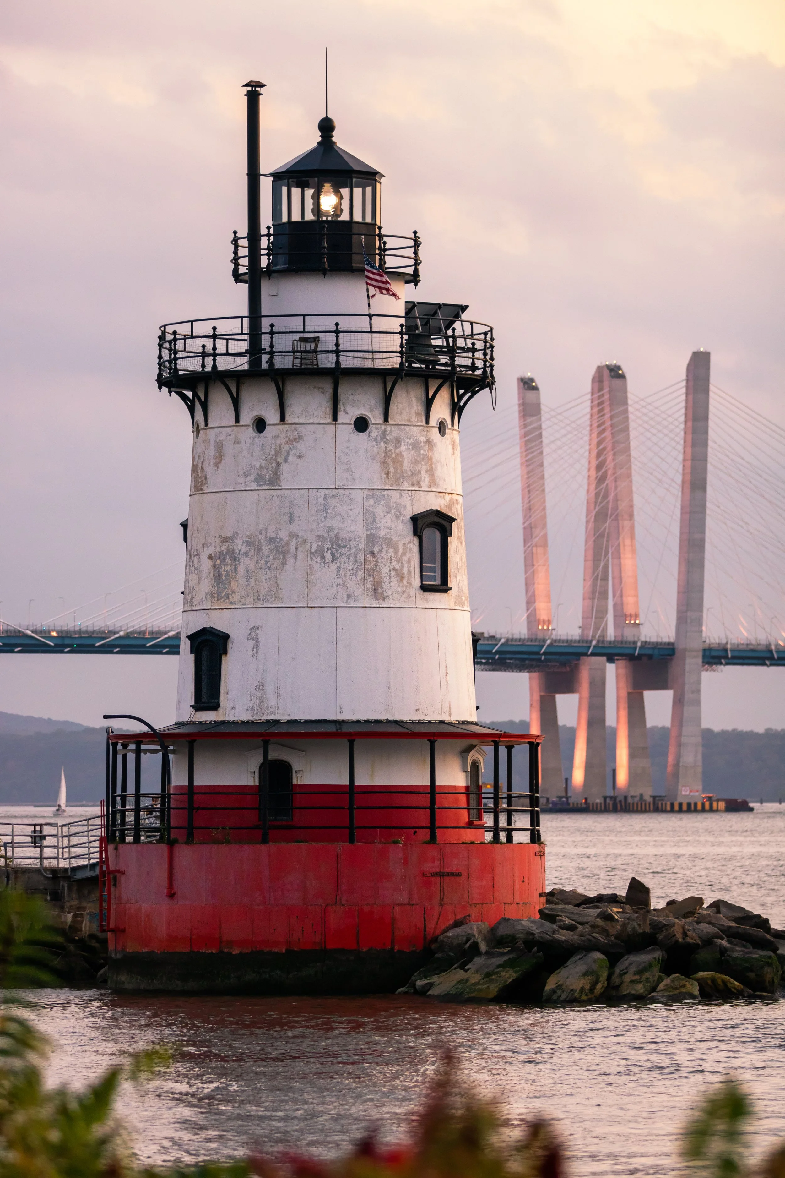 A lighthouse with a red base and black railing, standing on rocks near the water, with a bridge featuring tall, illuminated pylons in the background.