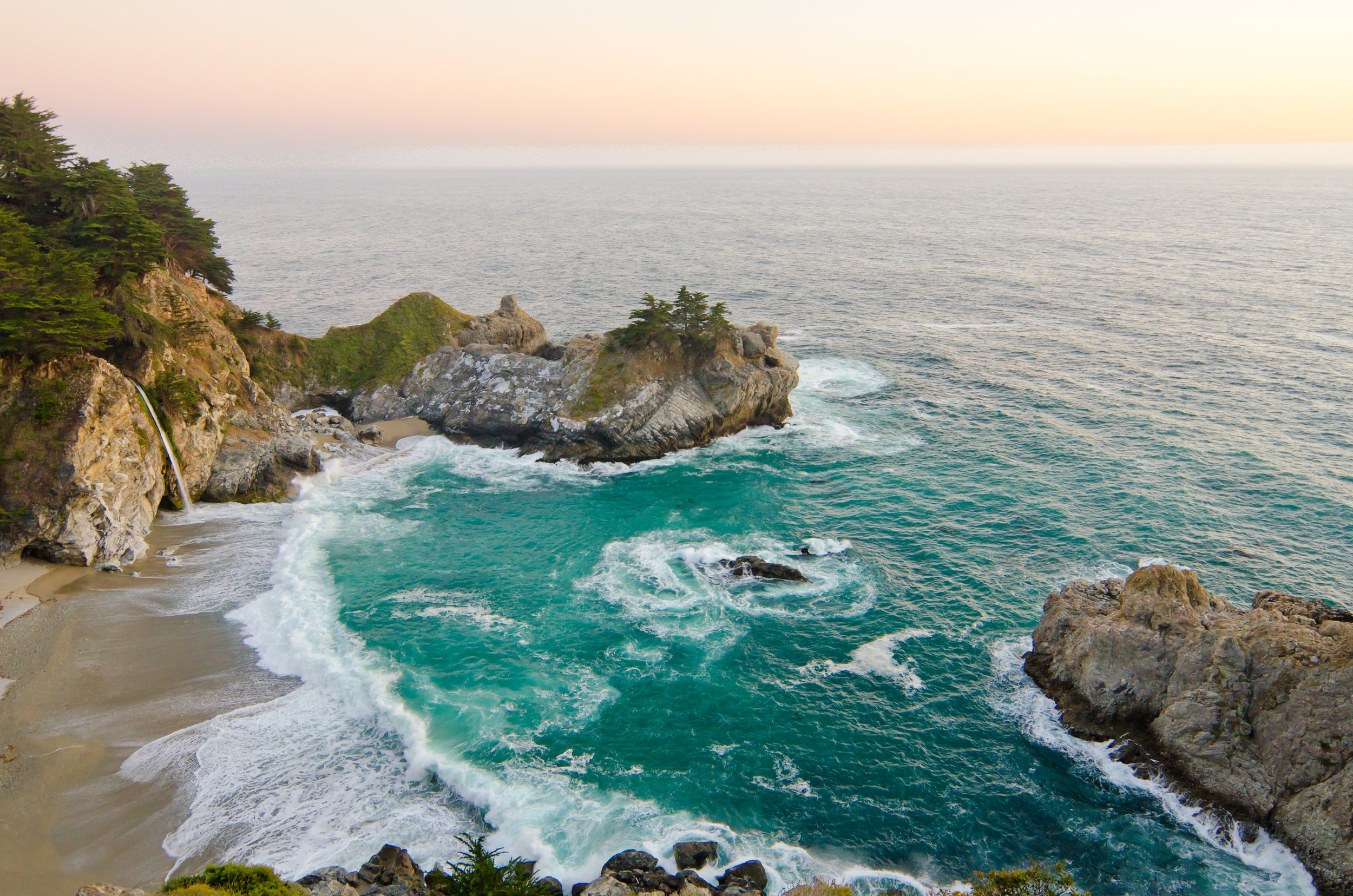 A rocky coastline with a small waterfall on the left, a sandy beach, and turquoise waves crashing against rocks, under a pastel-colored sky.