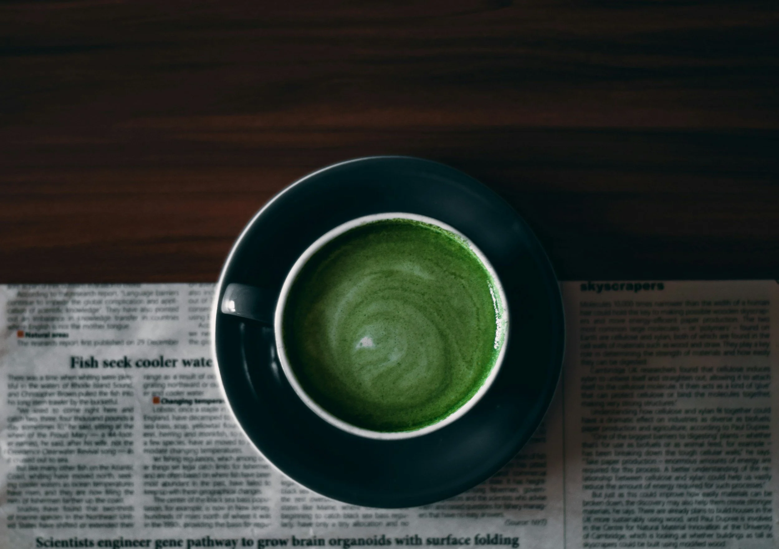 A top-down view of a black cup filled with green matcha tea on a black saucer, placed on a newspaper on a wooden surface.