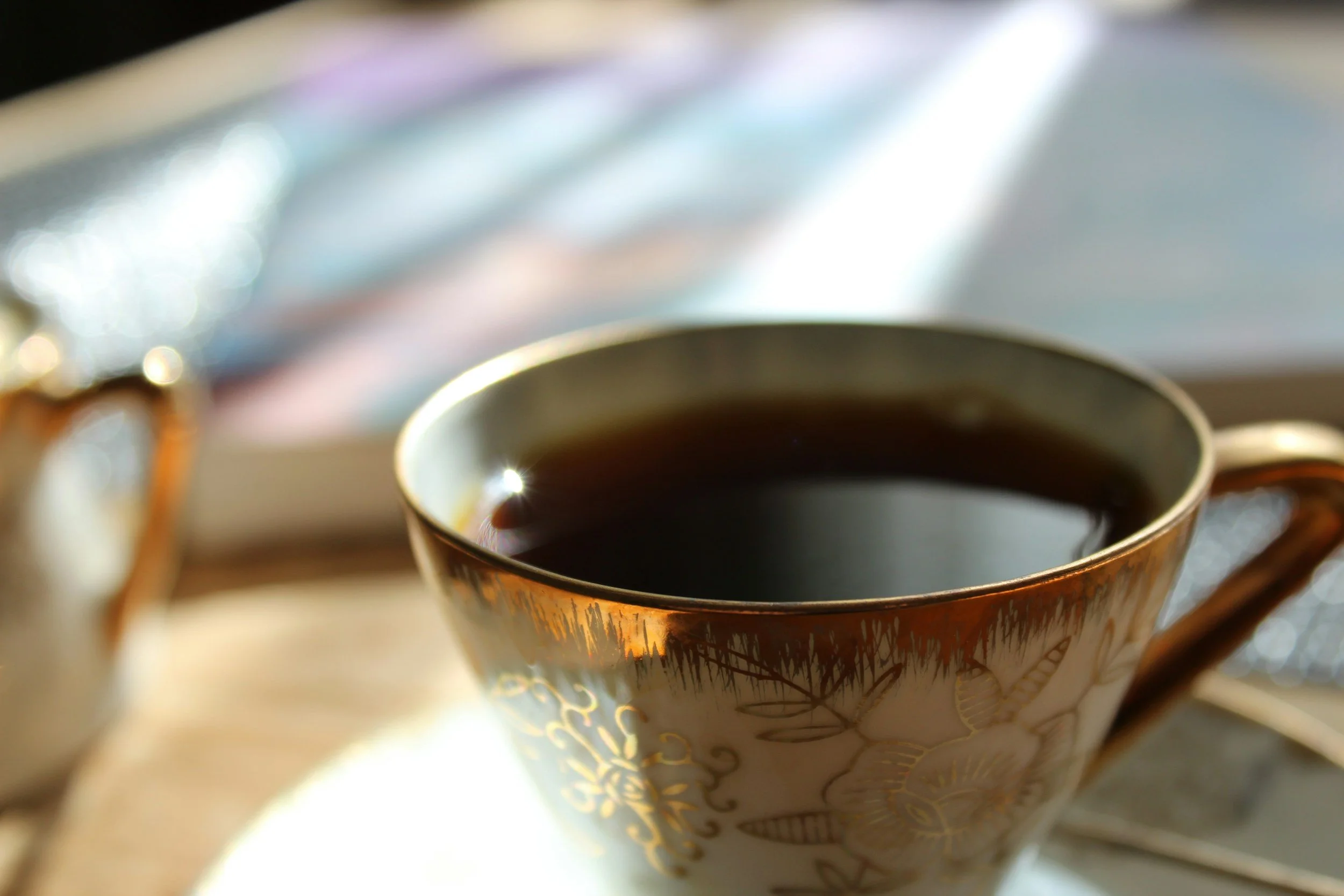 A close-up of a decorative teacup filled with black coffee, placed on a wooden surface with a blurred background including what appears to be a book and some colorful objects.