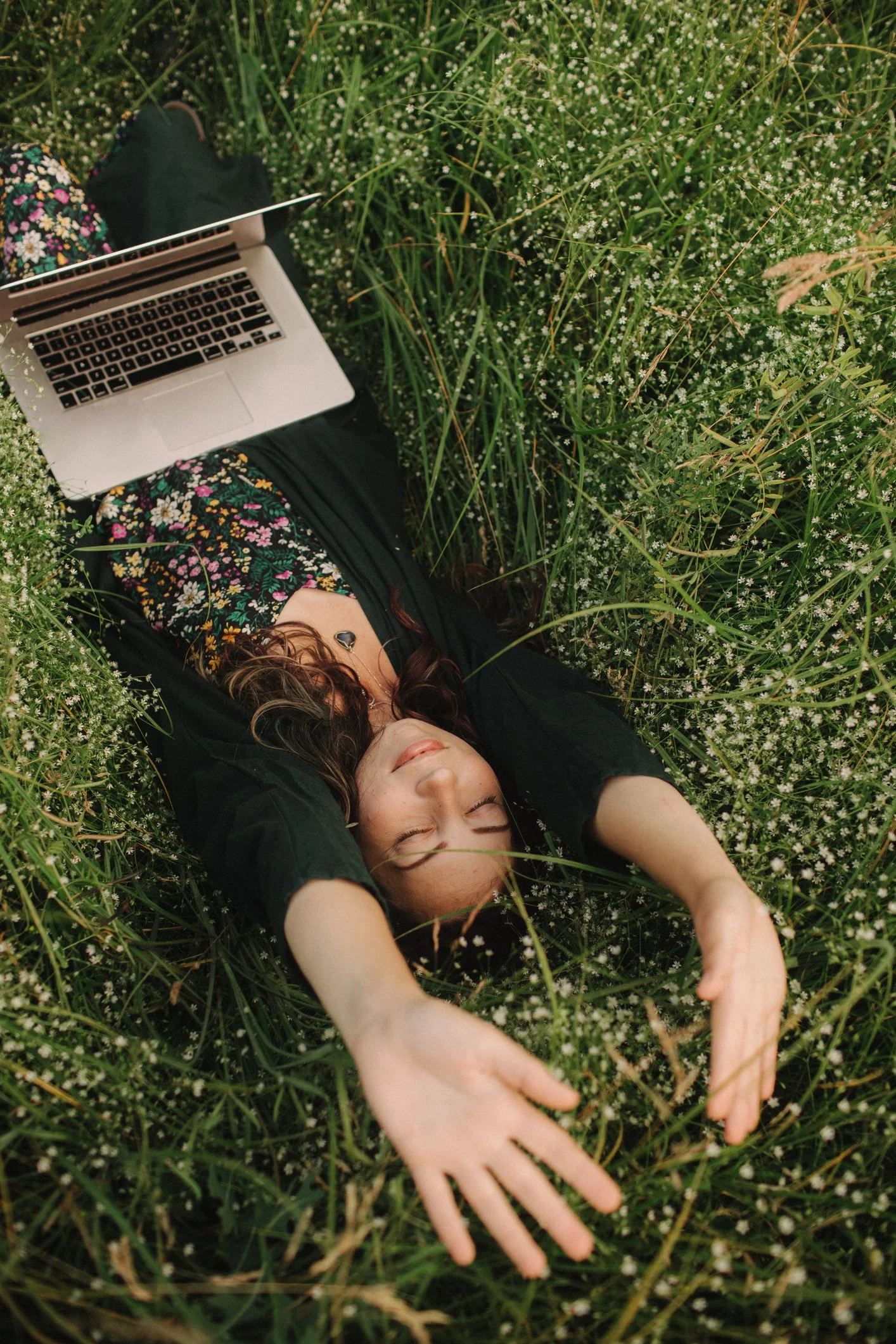 A woman wearing a floral dress and black jacket lying on her back in a field of small white flowers, with a laptop on her lap and her eyes closed.