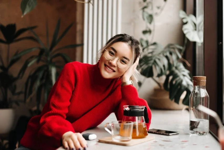 A woman in a red sweater with glasses smiling, sitting at a table with a glass teapot, a mug, a water bottle, and a smartphone, in a cozy indoor setting with plants.