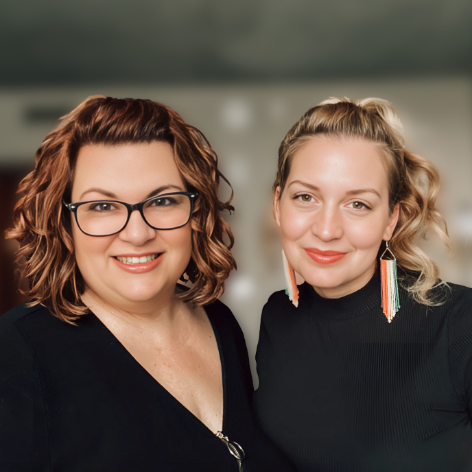 Two women smiling, one with glasses and curly hair, the other with wavy blonde hair and colorful earrings, both wearing black tops.