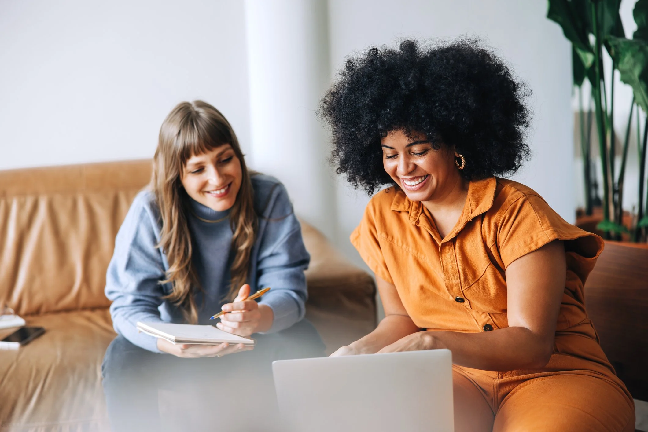 Two women sitting on a couch, looking at a laptop, smiling, with one holding a notebook and pen, in a cozy room with plants in the background.