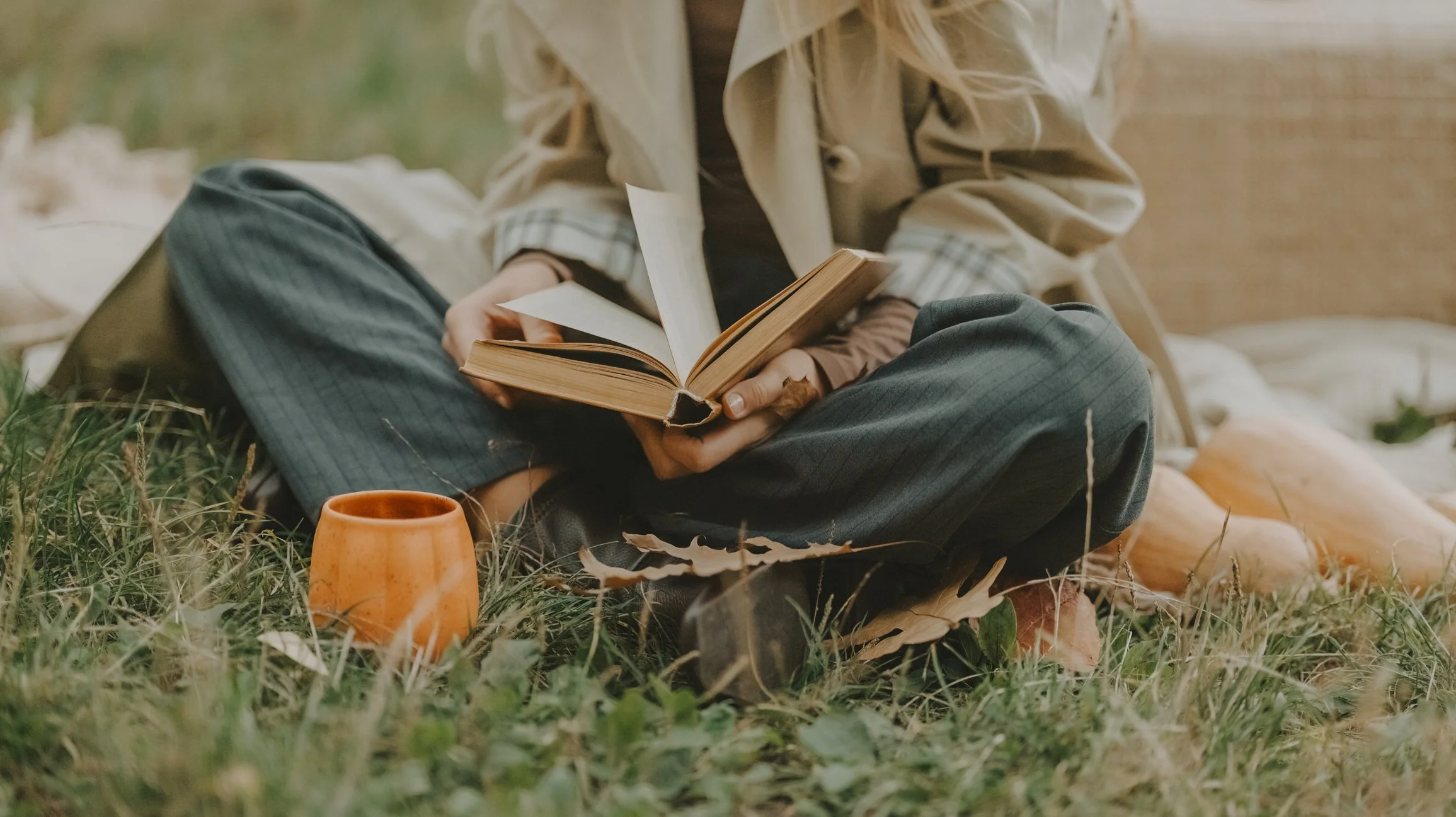 Person sitting outdoors on grass, reading a book, with a ceramic mug nearby.
