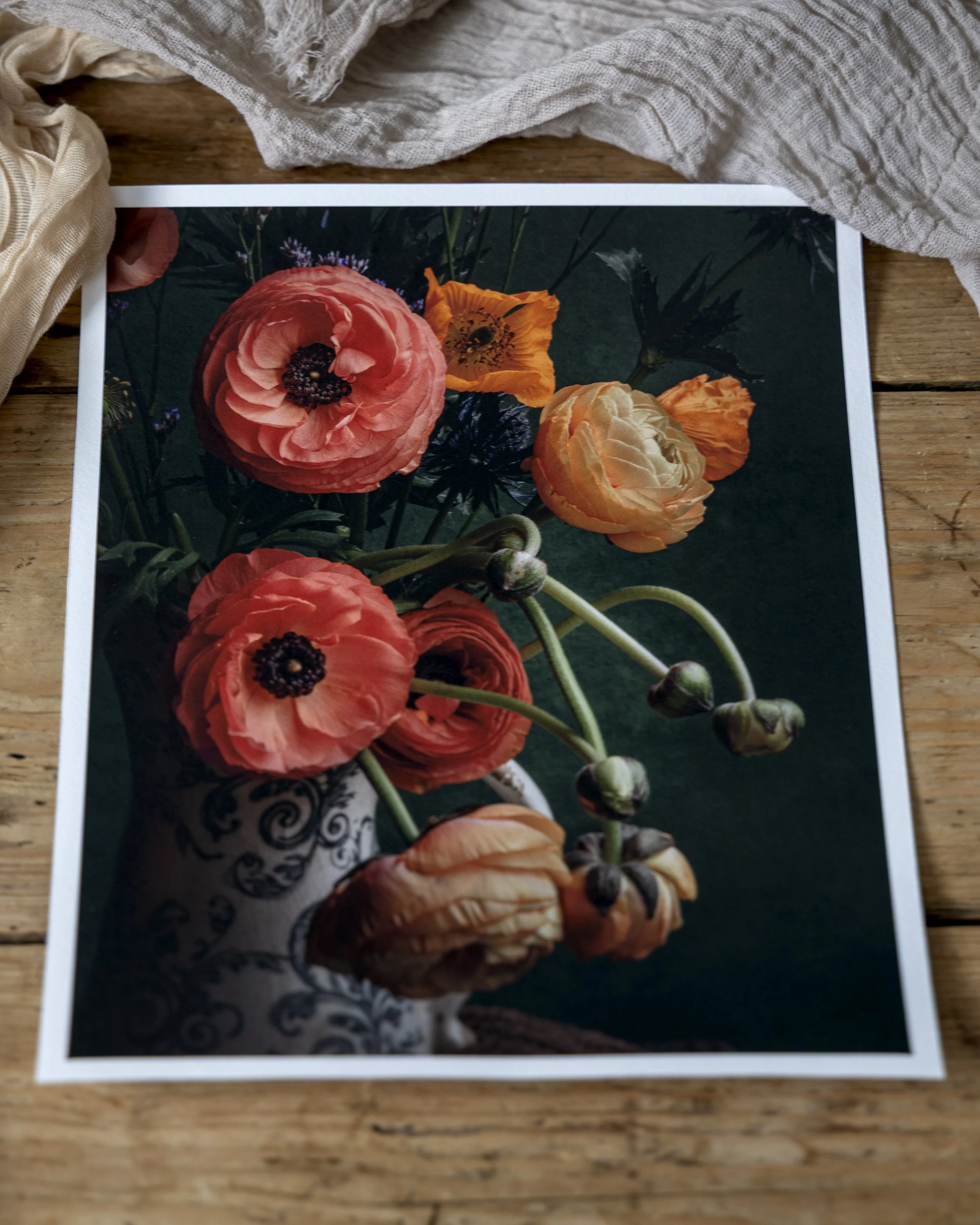 A printed photograph of colorful flowers, including pink and orange poppies, resting on a wooden surface with some cloth around it.