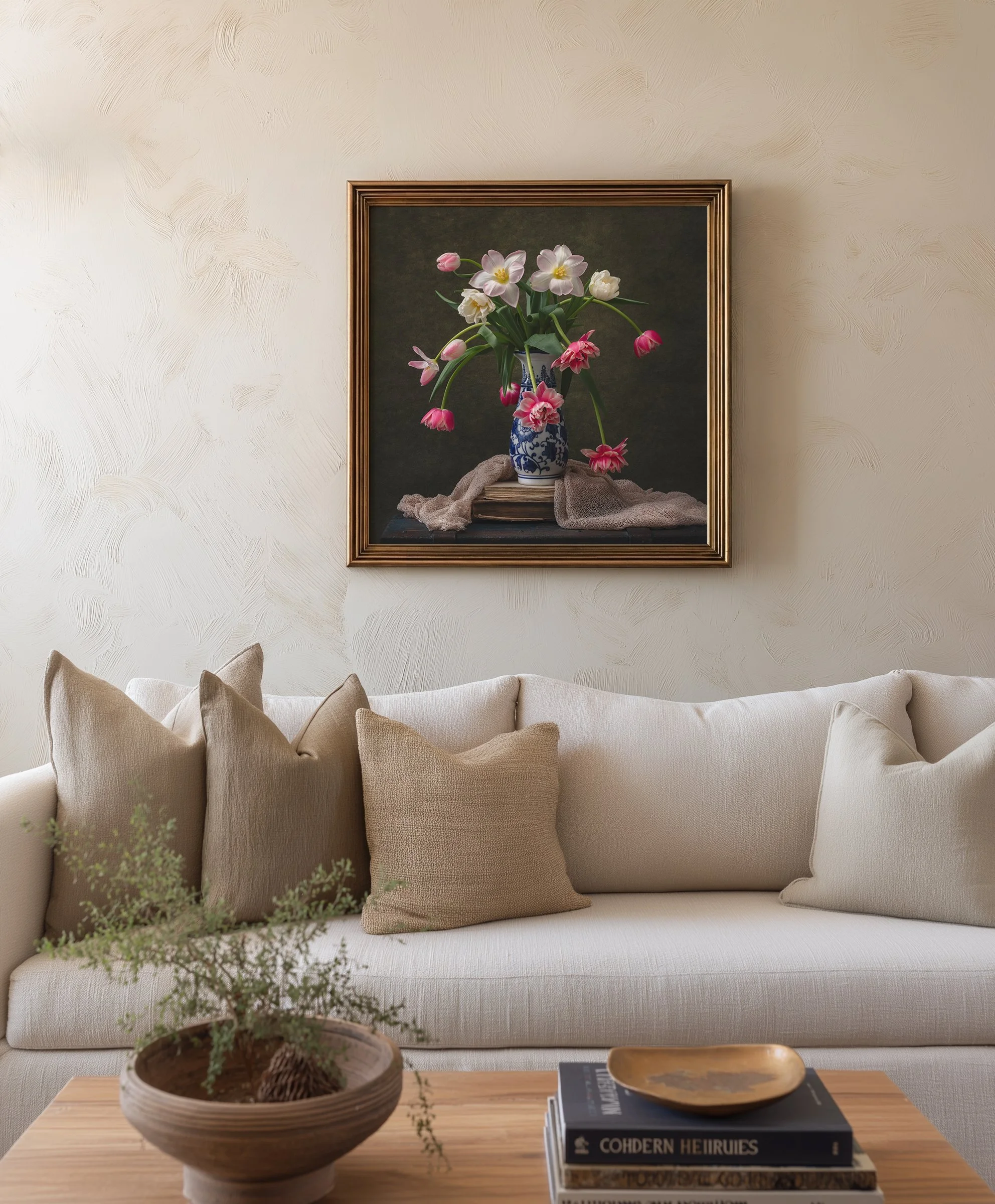 A cozy living room scene with a beige sofa decorated with beige cushions. In front of the sofa is a wooden coffee table holding a stack of books titled 'Cohdern Heiruis' and other titles, along with a decorative shallow bowl. To the left of the table, a potted shrub with green foliage is placed. On the wall behind the sofa, a framed still life painting features a blue and white vase with a variety of pink and white flowers. The wall has a textured, off-white finish.