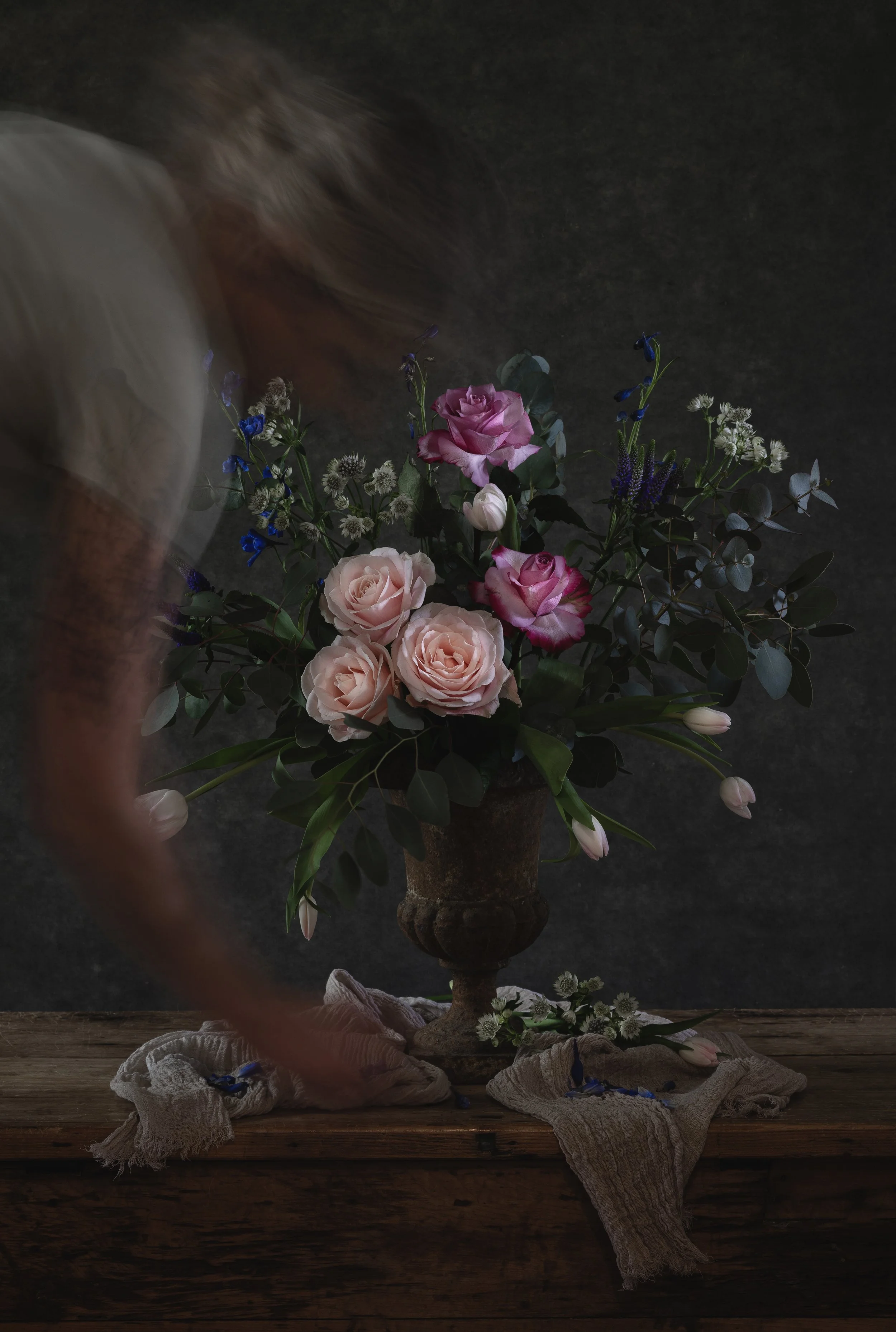 A floral arrangement featuring pink and cream roses, blue and white smaller flowers, and green foliage in a dark vase on a wooden surface with cloths and flowers around the base, against a dark background.