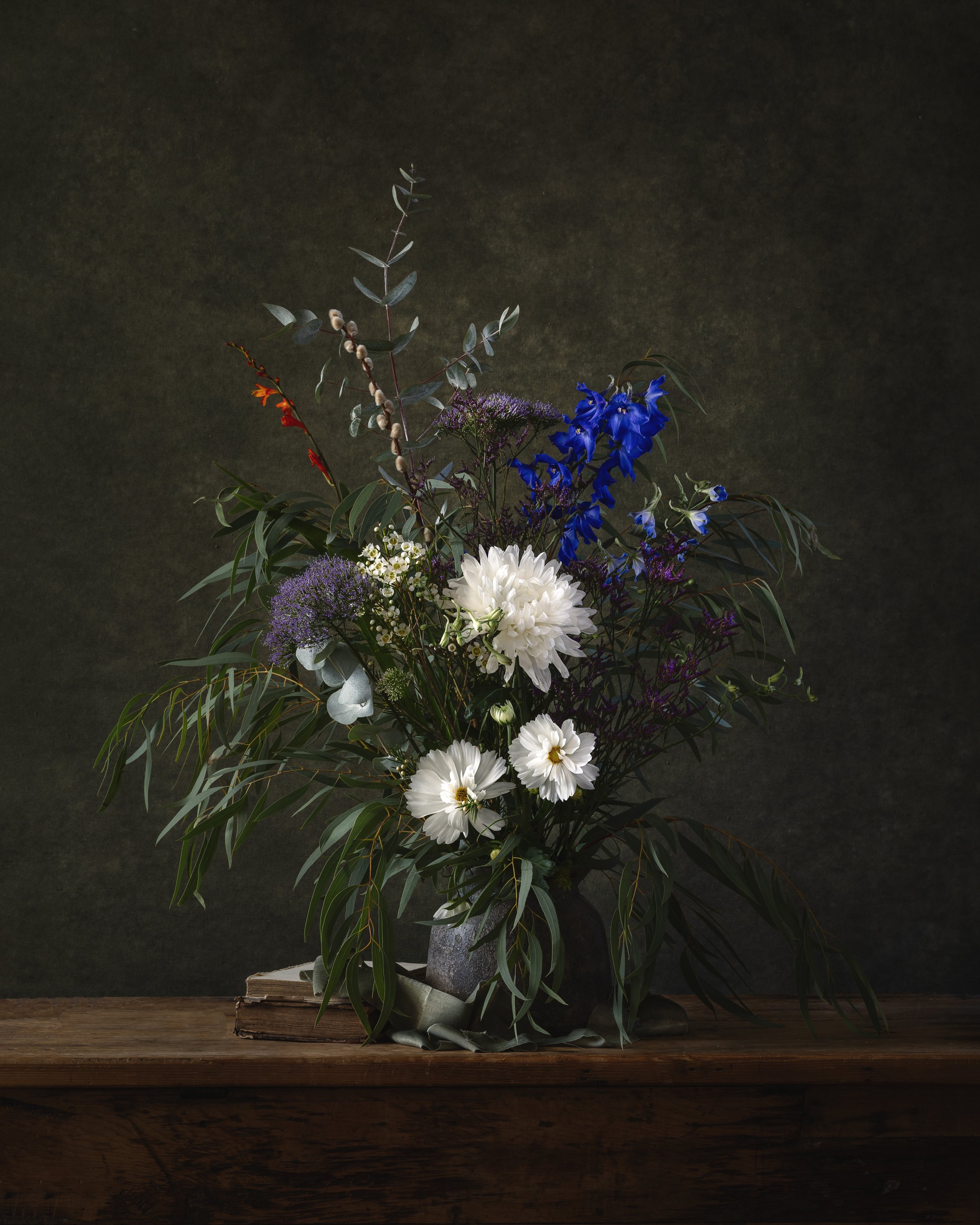 A floral arrangement with white, purple, and blue flowers on a wooden table, against a dark background.