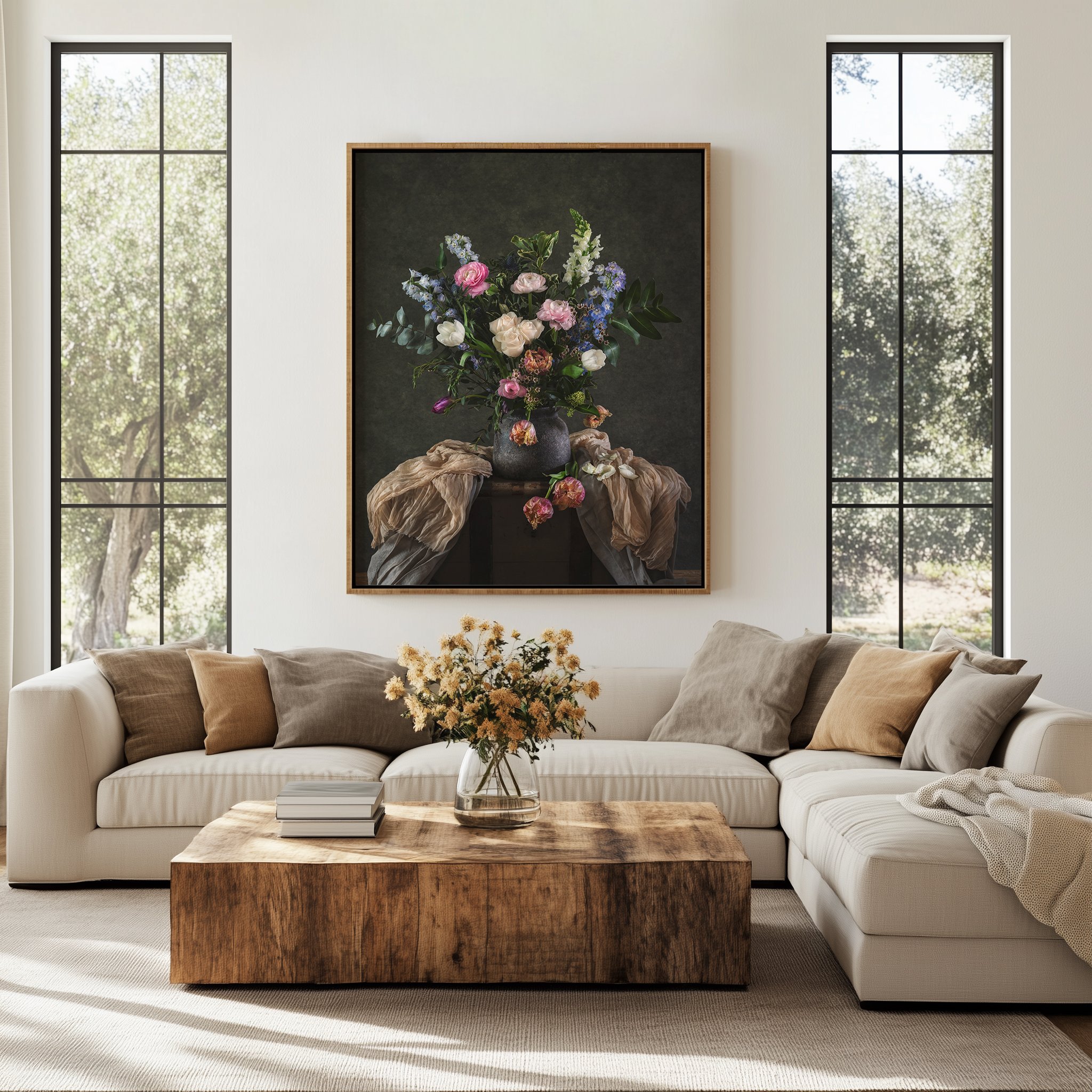 A living room with a white sectional sofa, beige and gray pillows, a wooden coffee table with a glass vase of yellow flowers, a framed floral painting on the wall, tall windows with black frames letting in sunlight, and a view of trees outside.