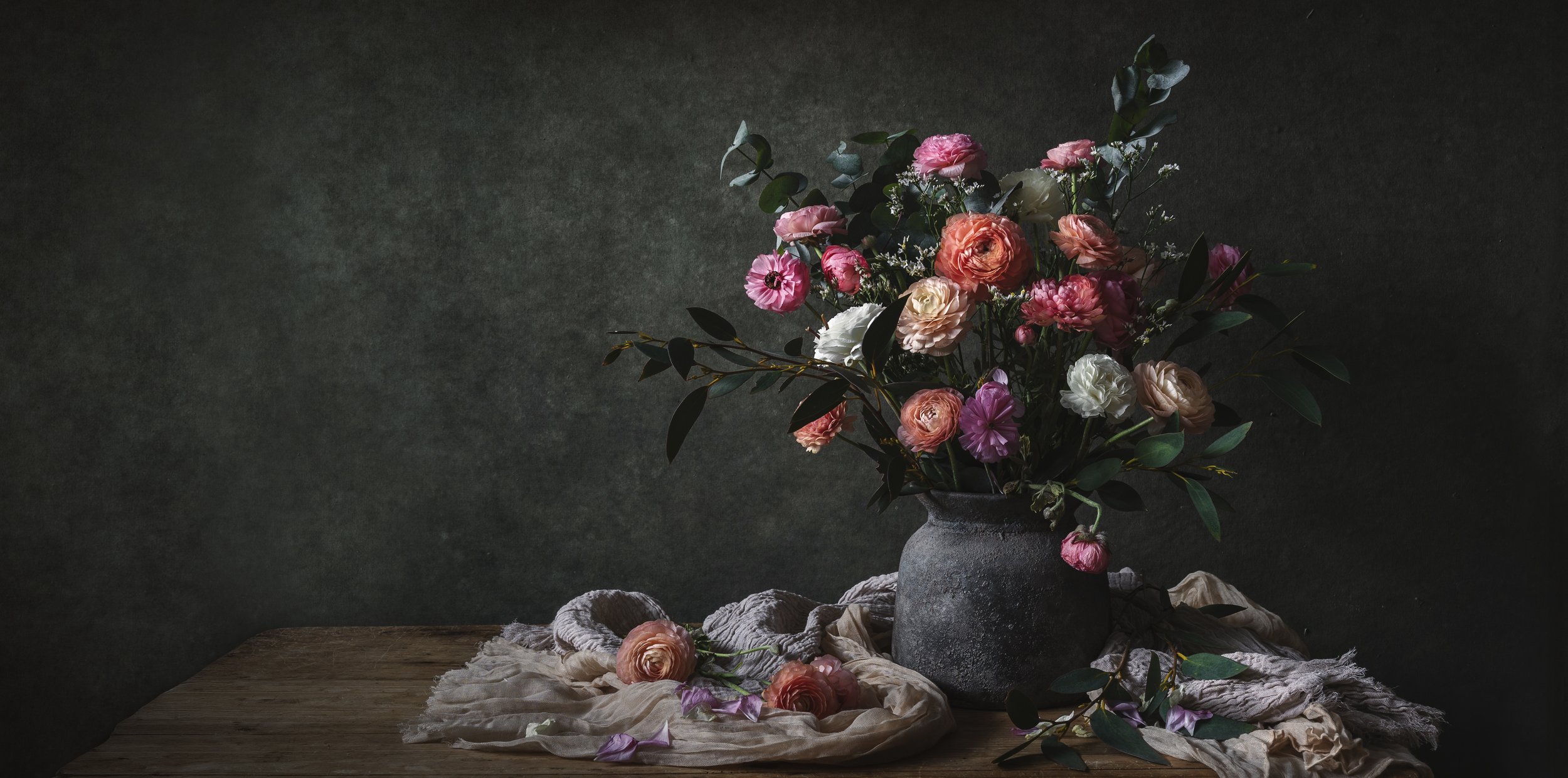 A bouquet of pink, white, and peach flowers with green leaves in a gray textured vase on a wooden table with soft draped cloth and scattered flowers, against a dark textured background.