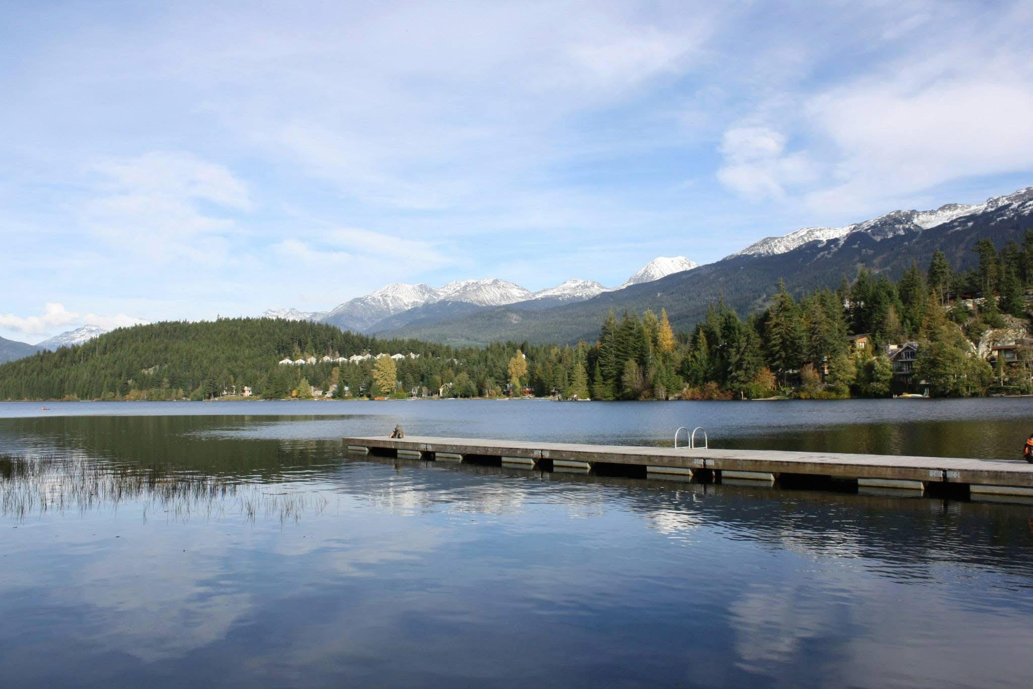 A peaceful lake surrounded by forested hills and snow-capped mountains in the distance under a blue sky with scattered clouds.