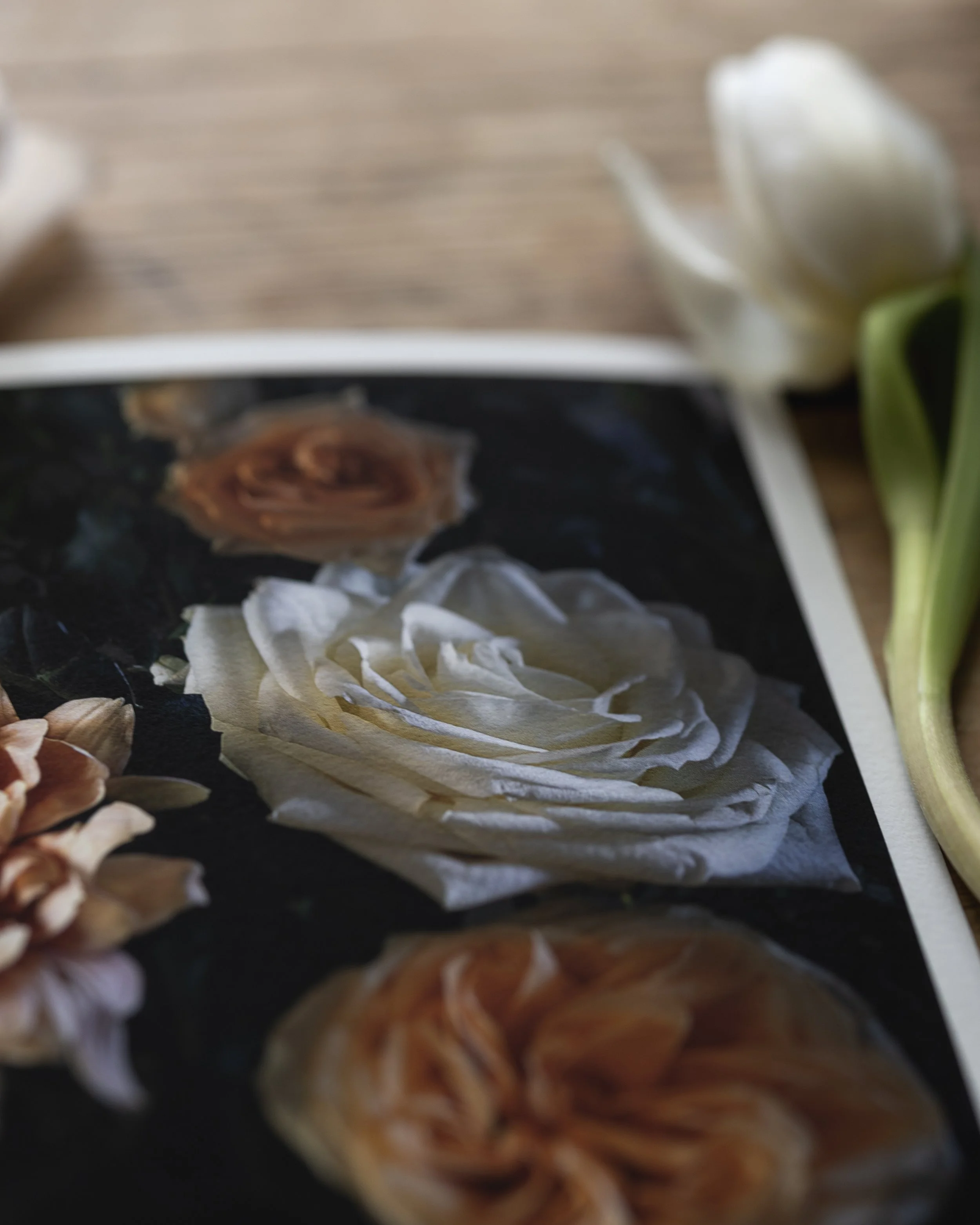 Close-up of printed floral photographs showing various flowers, such as roses and peonies, with a white tulip and some other flowers partially visible around the print on a wooden surface.