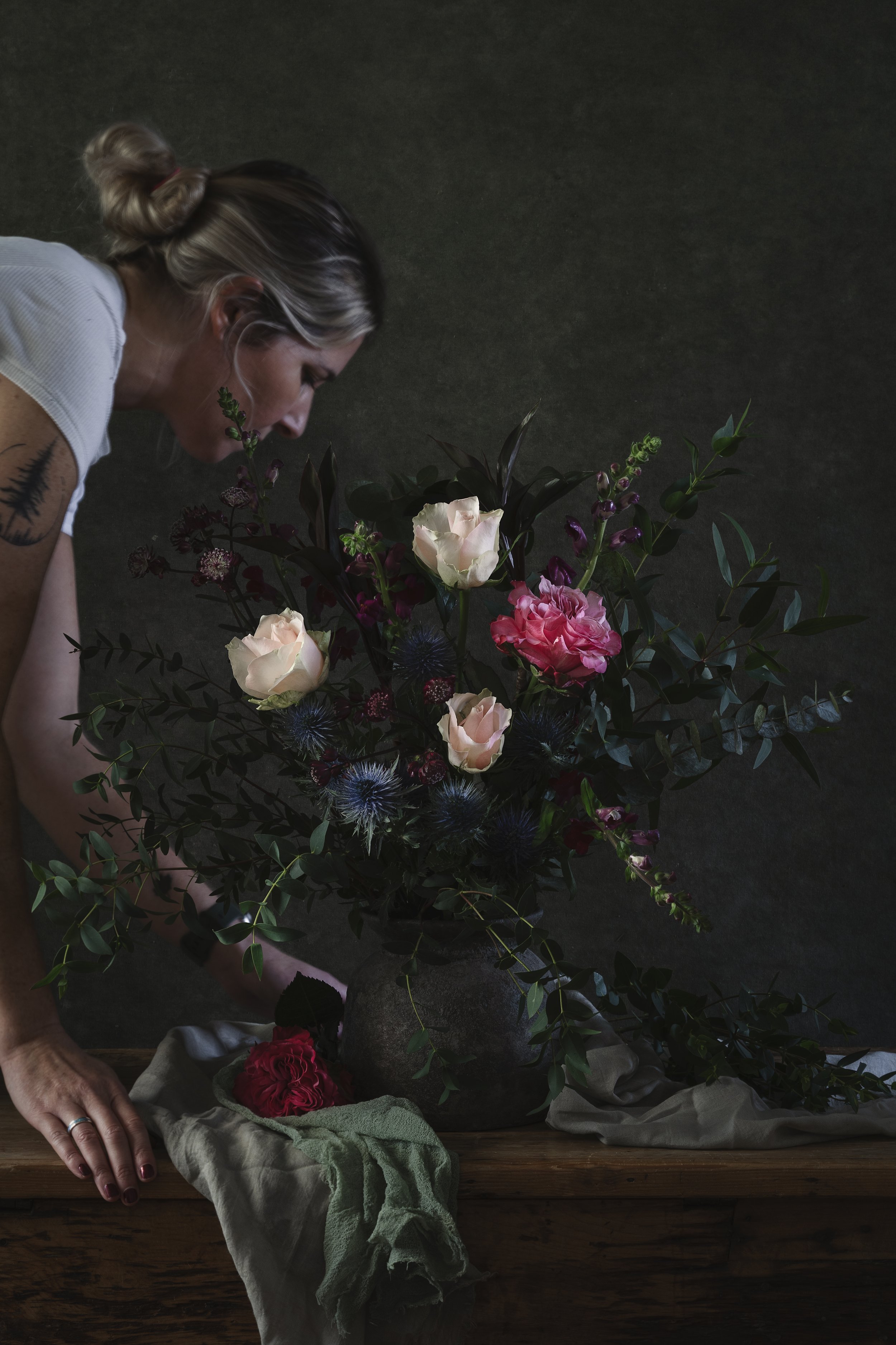 A woman with blonde hair styled in a bun arranging a large mixed flower bouquet on a wooden table.