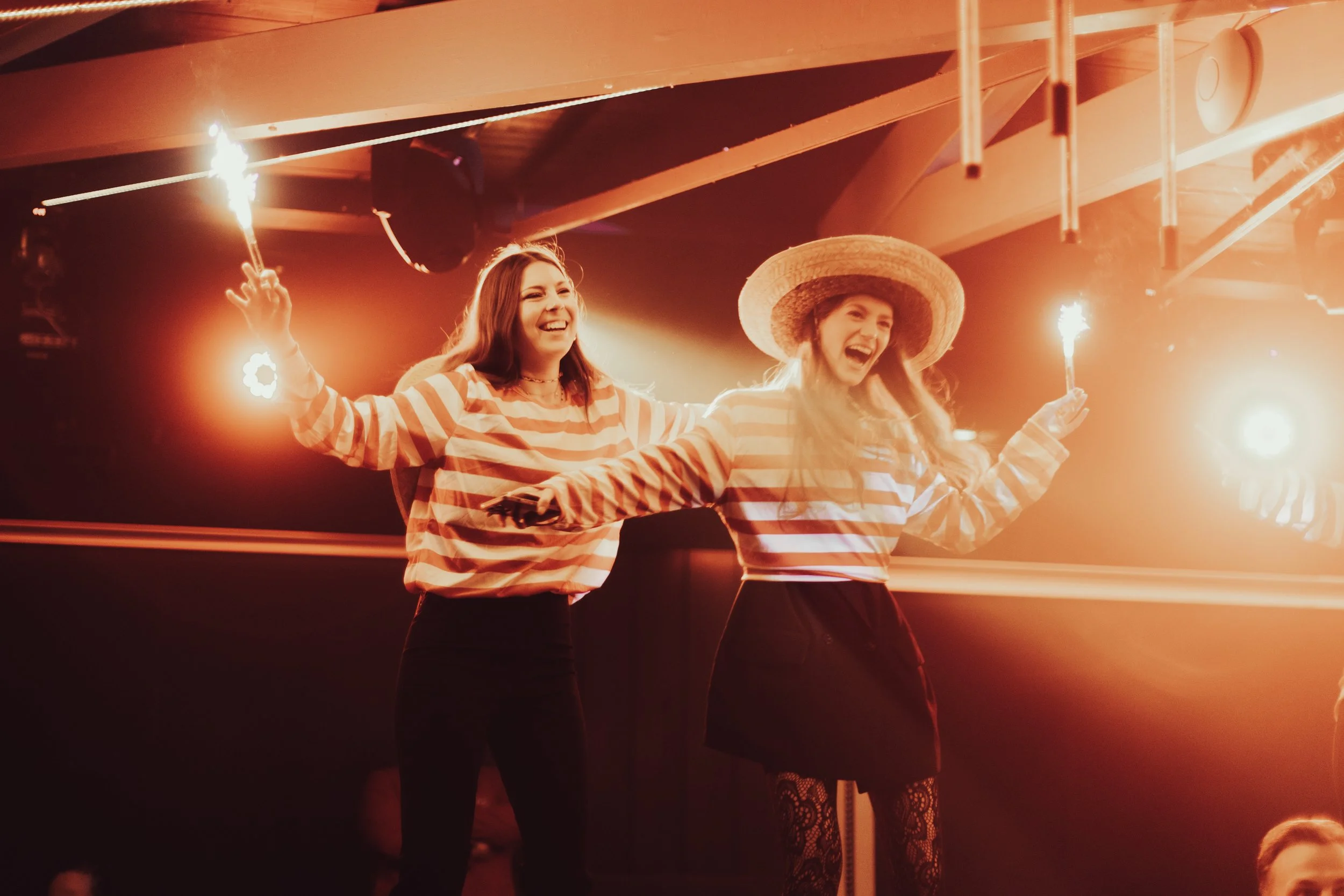 Deux femmes souriantes portent des chemises à rayures et décorent un décor festif, tenant des cierges magiques allumés. L'une porte un chapeau de paille, toutes deux semblent célébrer quelque chose dans une ambiance chaude et lumineuse.