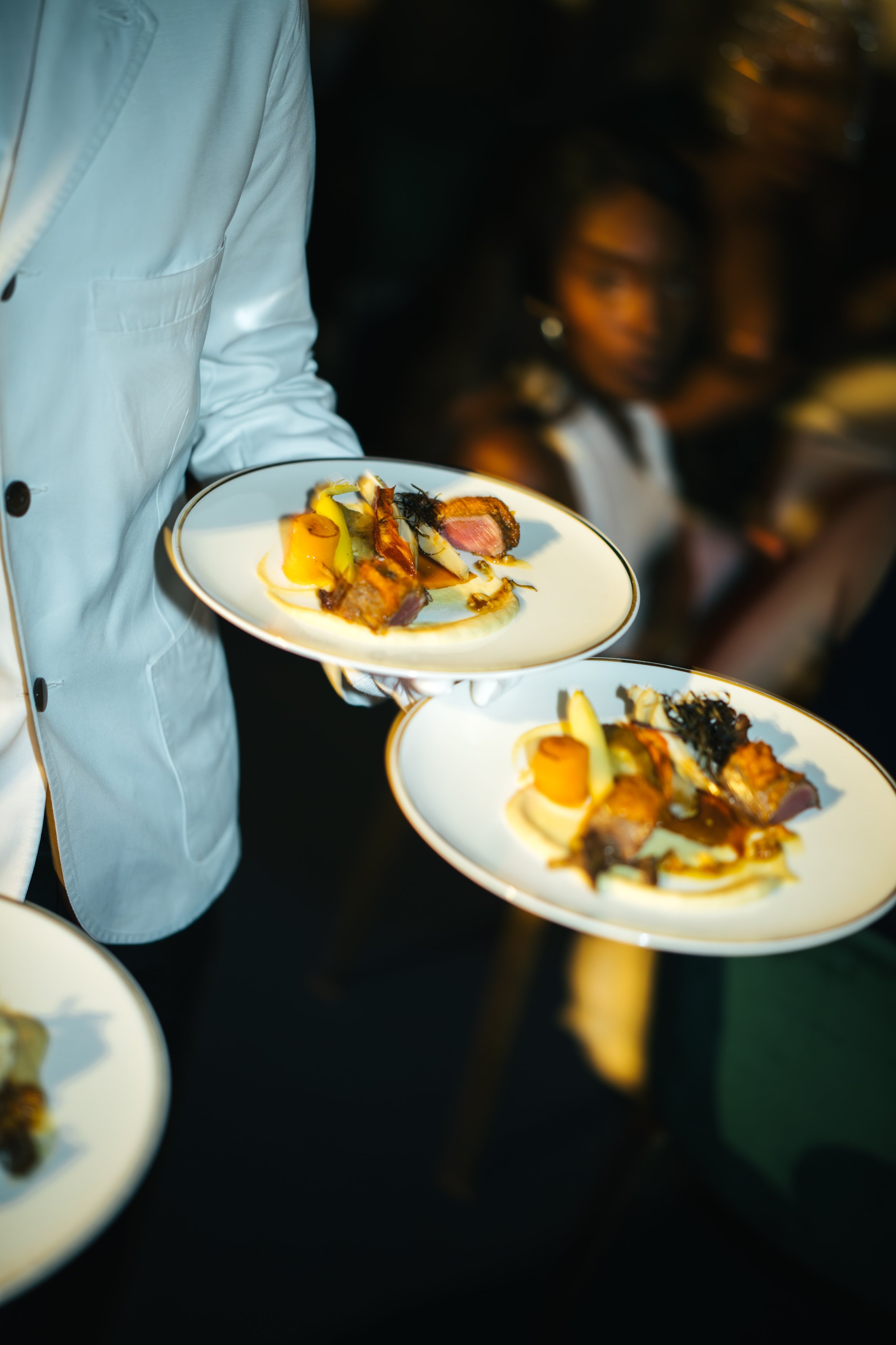 Serveur en veste blanche tenant deux assiettes avec des plats gastronomiques, autre personne floue en arrière-plan