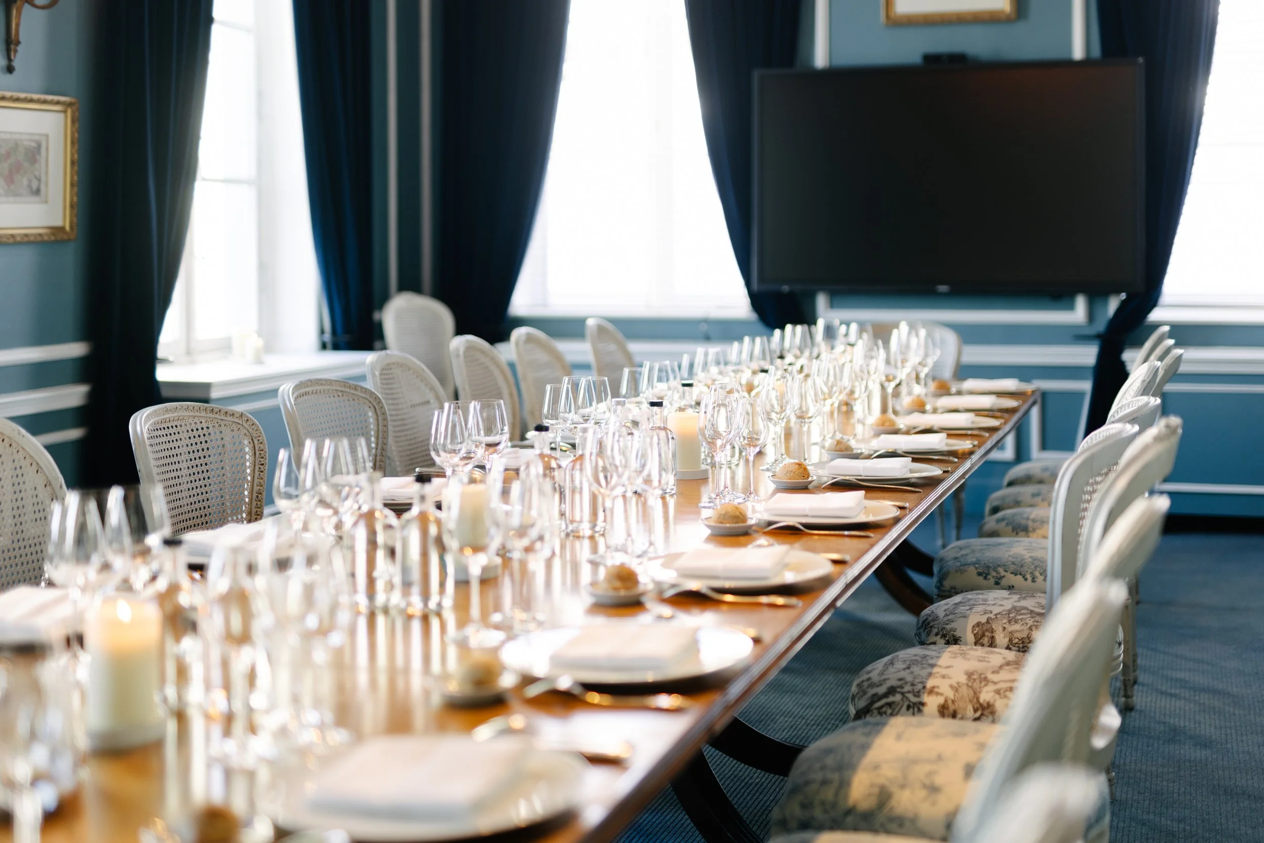 Salle de réunion élégante avec une longue table, décorée avec des verres à vin, assiettes, bougies et couverts, entourée de chaises en bois avec assises rembourrées, grande télévision à l'arrière et rideaux noirs sur les fenêtres.