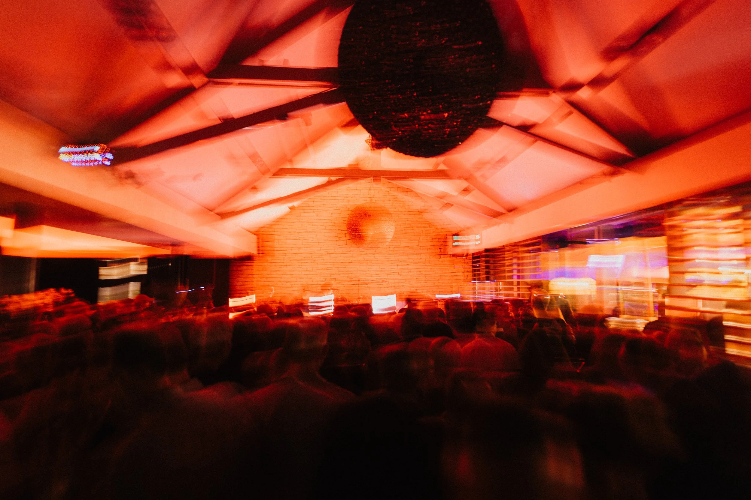 Une salle bondée avec un public regardant un spectacle ou une présentation dans un éclairage tamisé rouge. La scène est au centre, avec un mur en briques en arrière-plan.