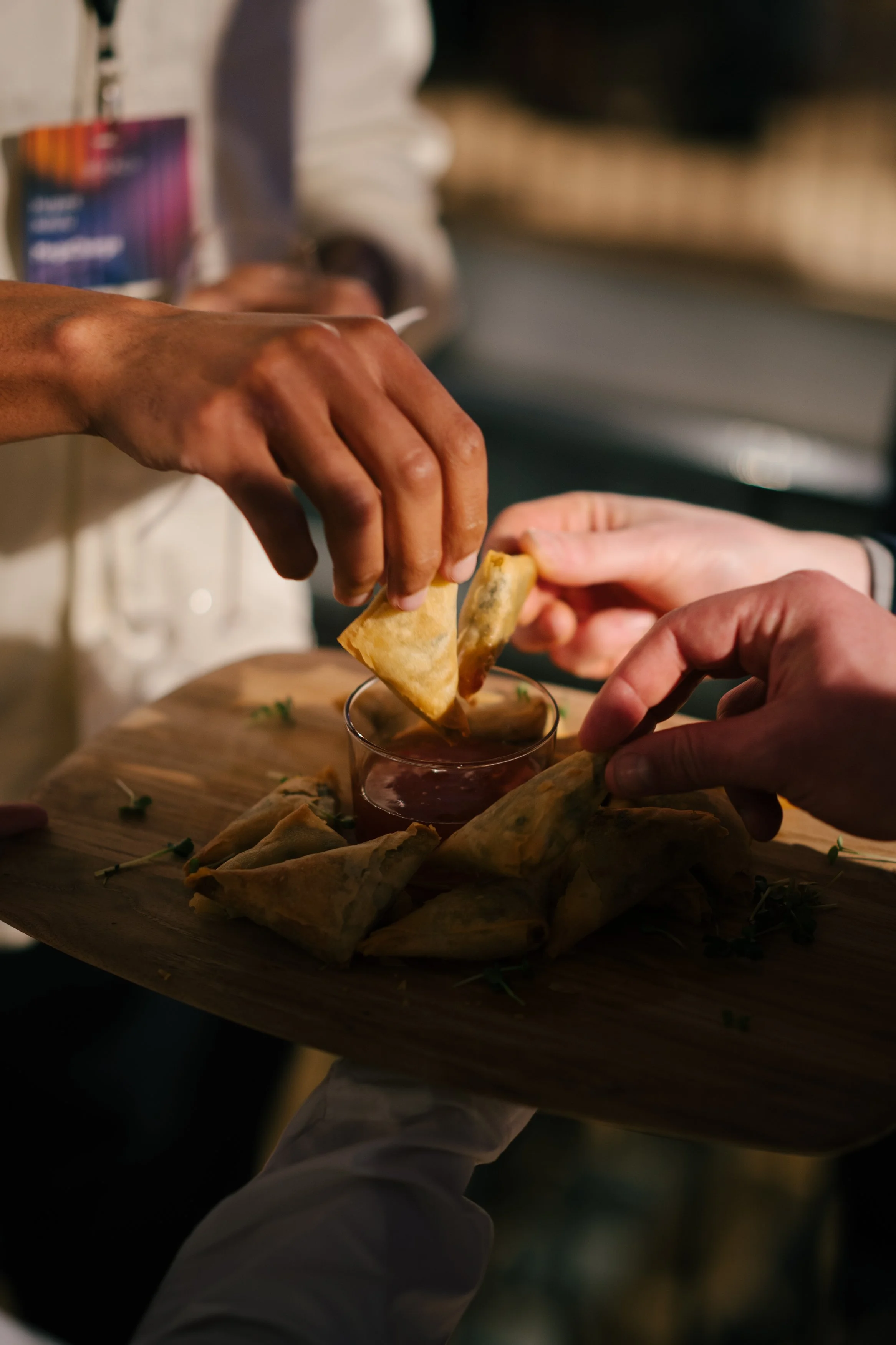 Deux mains, dont une appartient à une personne de peau foncée et l'autre à une personne de peau claire, échangent des rouleaux de printemps avec une sauce dans un verre en verre. Il y a d'autres rouleaux de printemps sur une planche en bois.