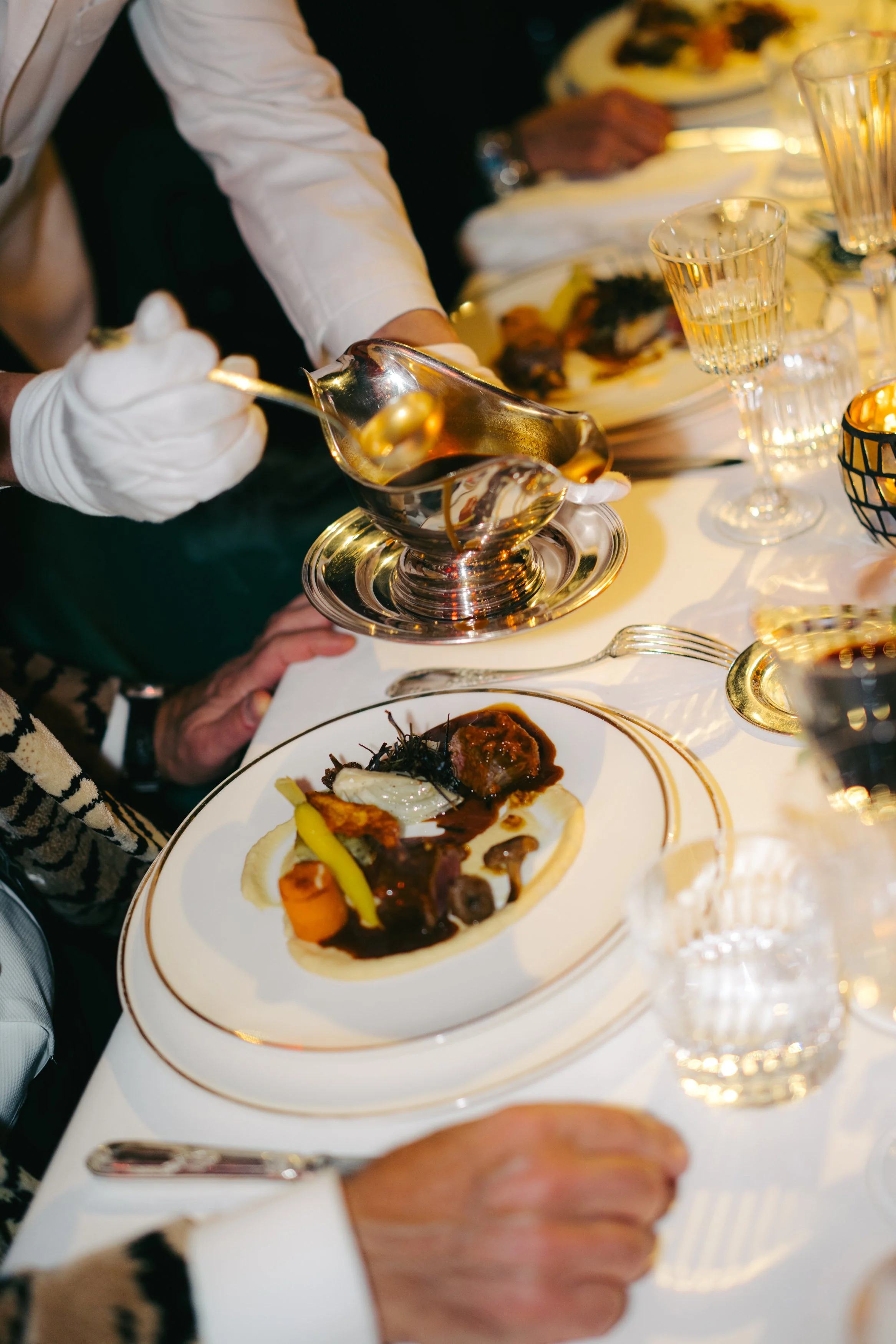 Une personne verse une sauce sur un plat étoilé dans une réception élégante avec un service en argent, des verres à vin et des plats de viande et légumes.