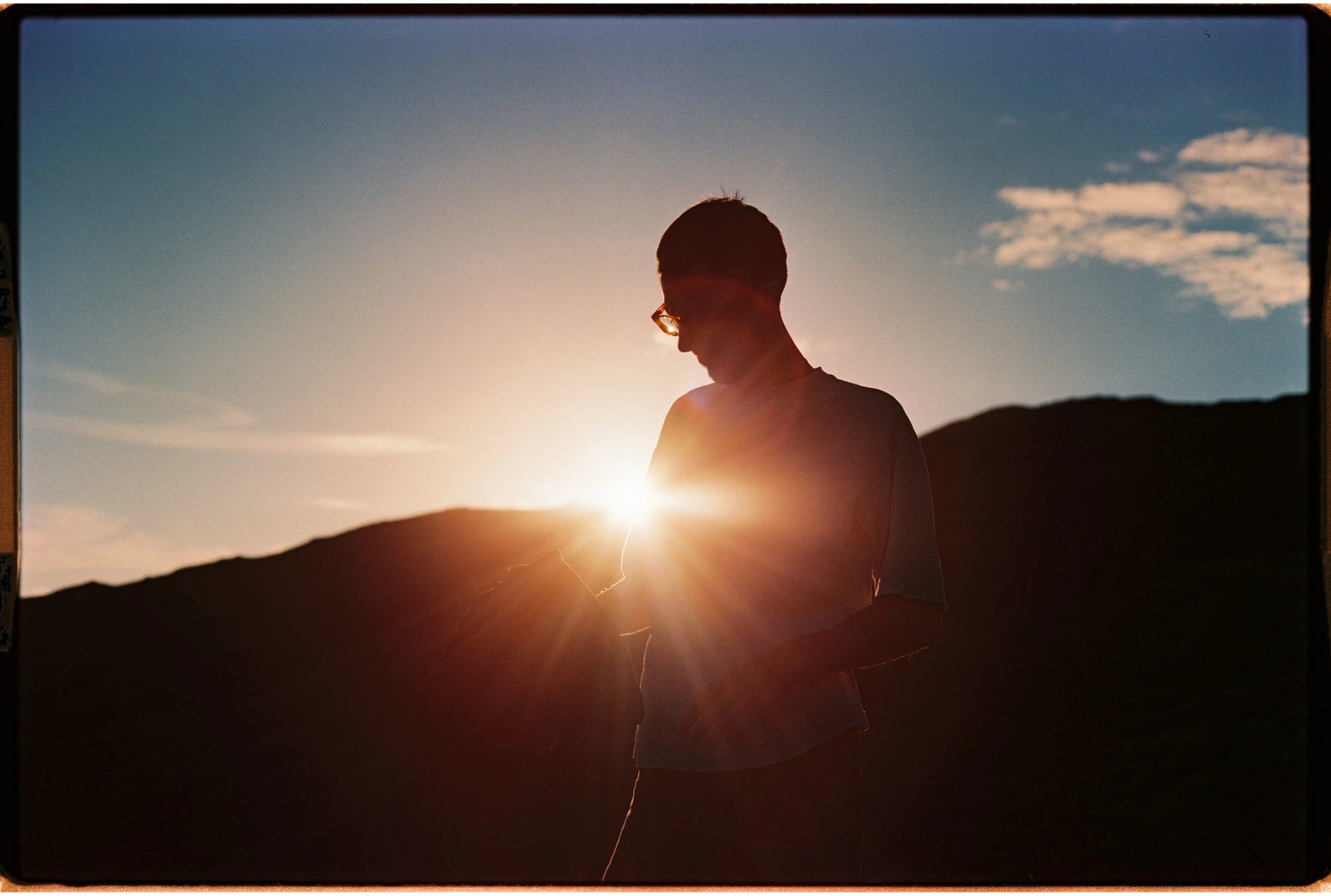 Un jeune homme en silhouette, portant des lunettes, regardant en bas alors que le soleil se couche derrière une montagne, créant une ambiance chaleureuse et sereine.