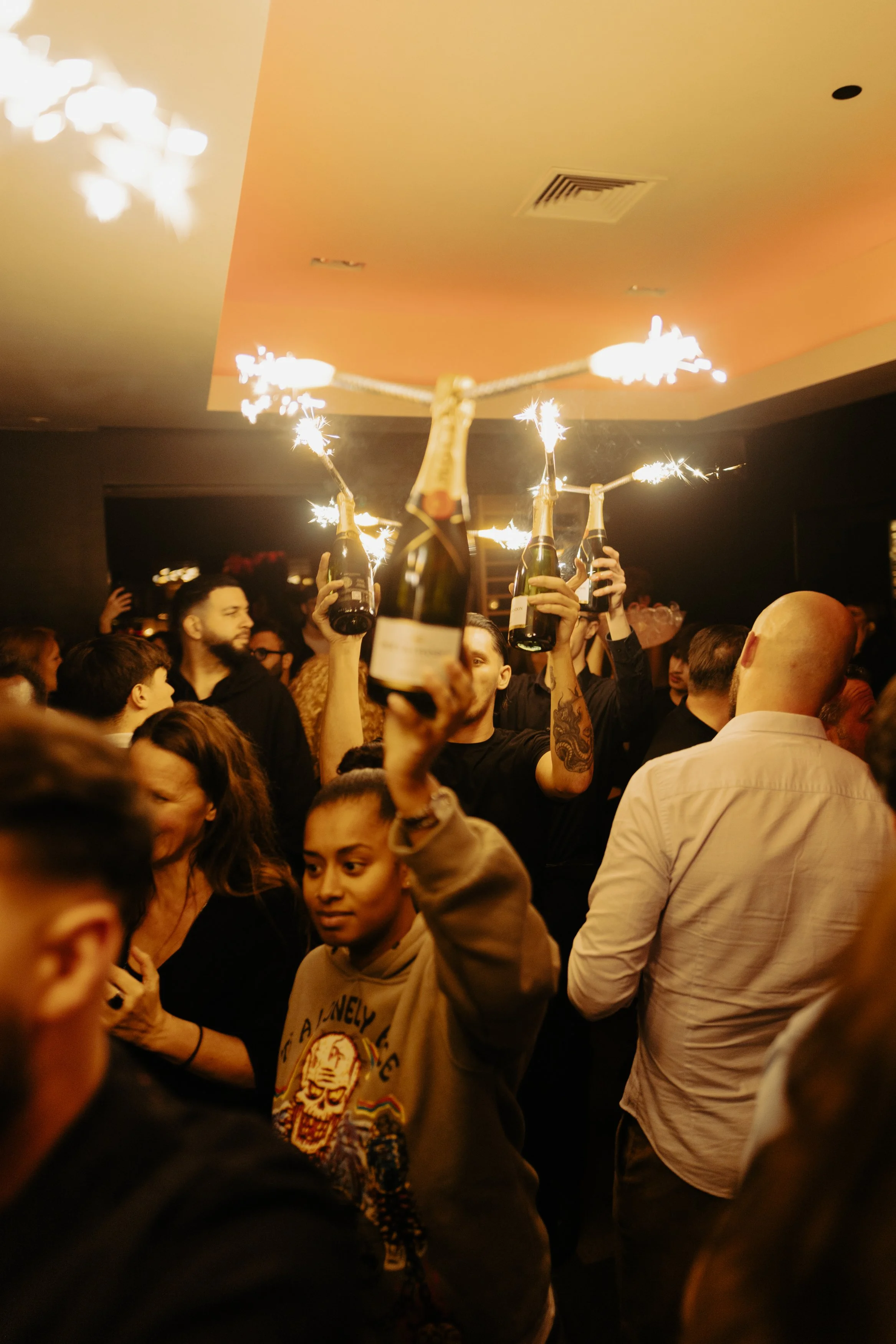 Groupe de personnes dans une fête ou un bar, certains tenant des bouteilles de champagne allumées avec des feux d'artifice.