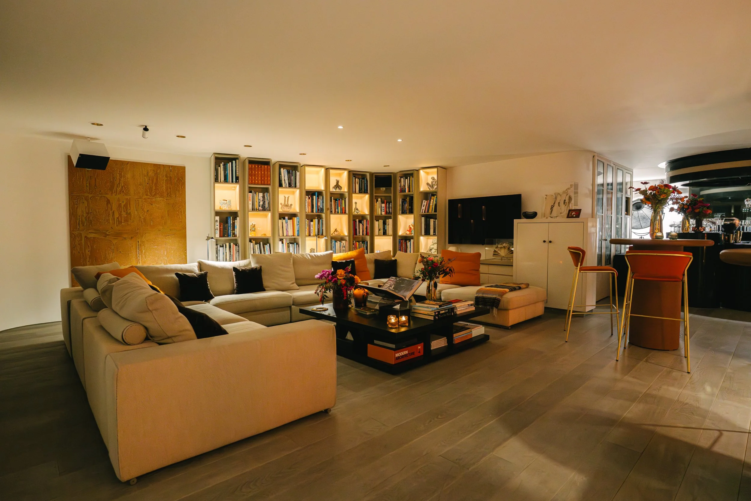 Salon moderne avec canapé beige, table basse, étagères avec livres, et décoration florale.