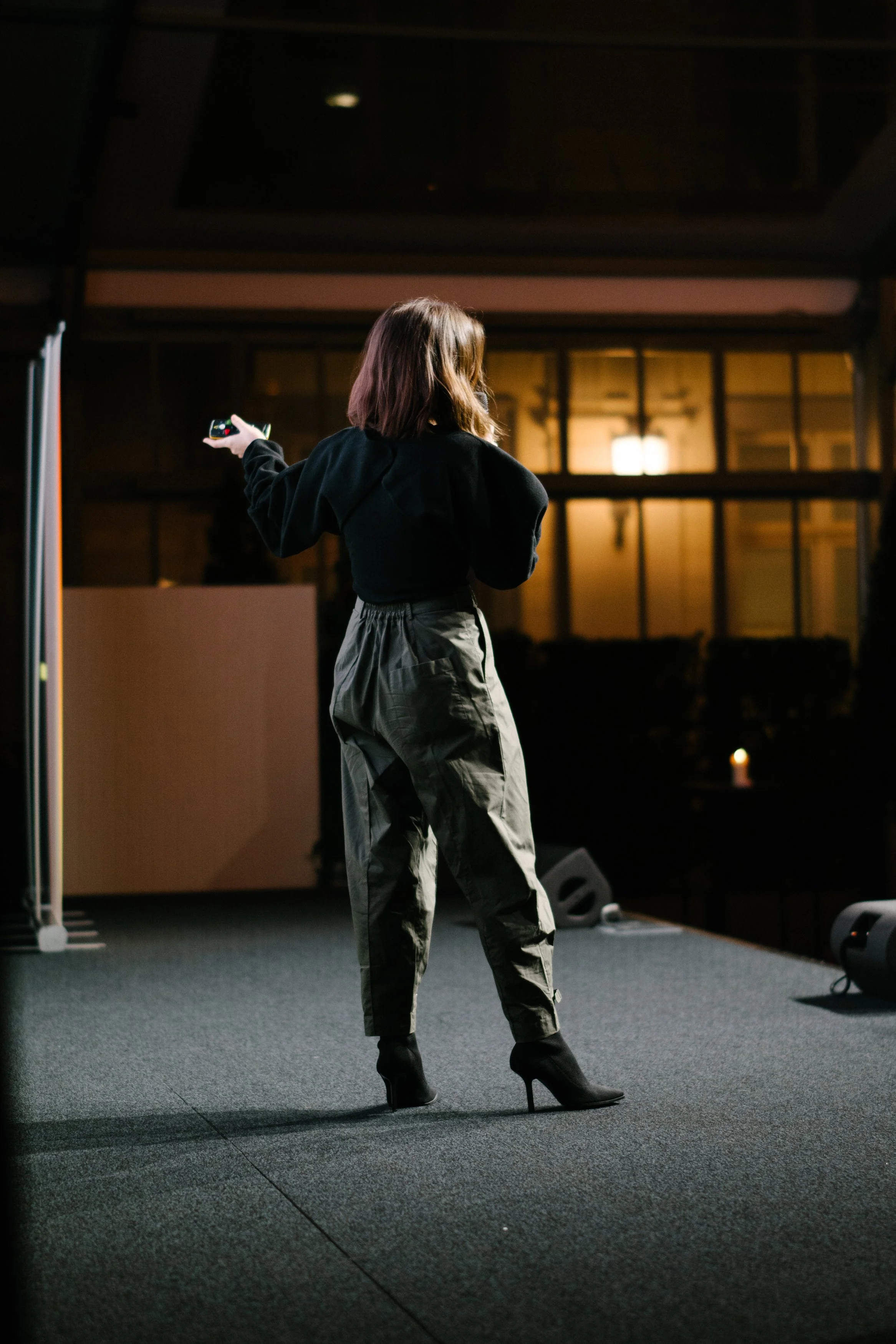 Une femme debout sur scène, de dos, tenant un microphone ou un objet dans la main, dans une pièce éclairée faiblement, probablement lors d'une présentation ou d'un spectacle.