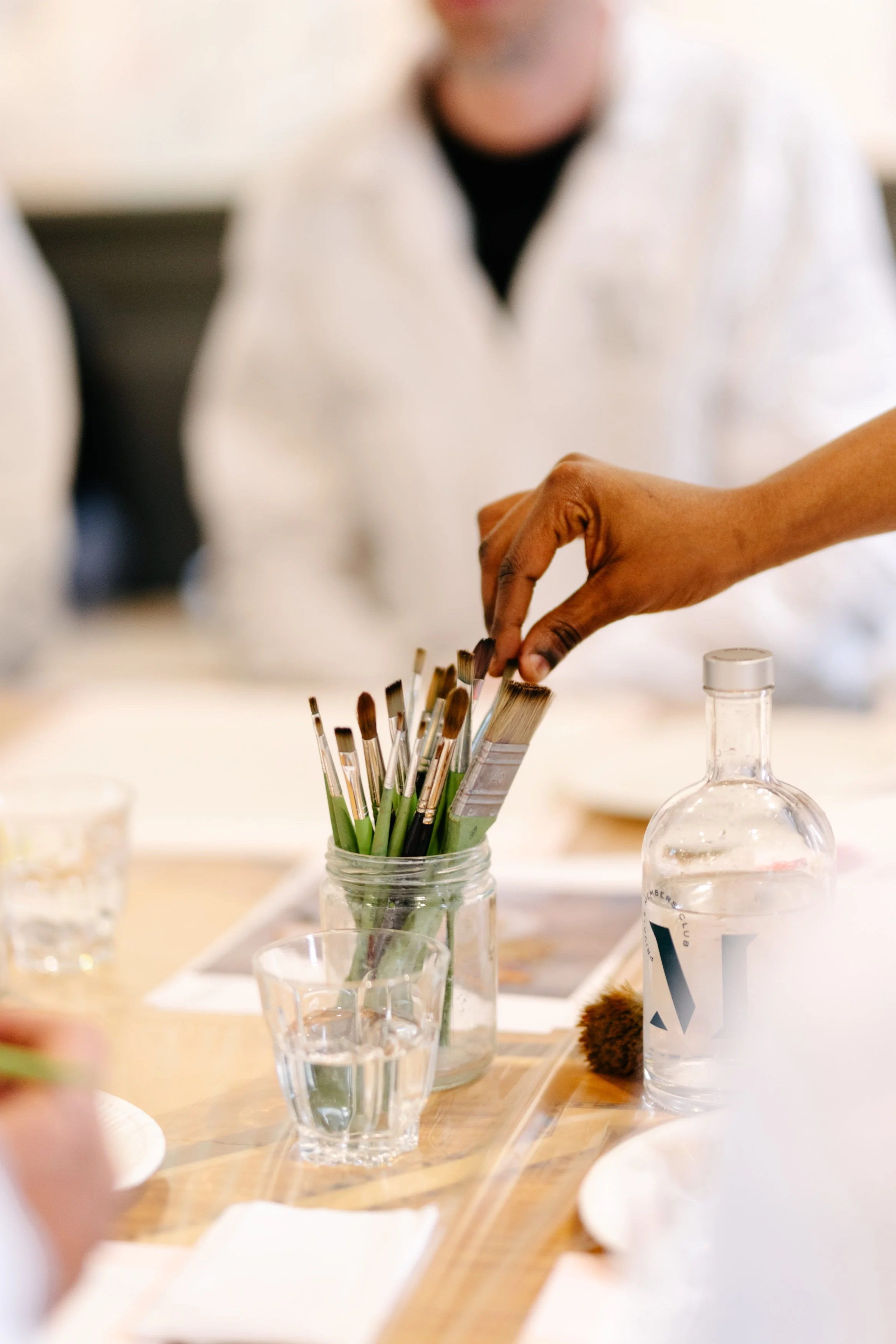 Mains d'une personne manipulant des pinceaux d'art dans un pot en verre, entourée de verres d'eau et d'une bouteille d'eau sur une table en bois, lors d'une activité artistique ou de peinture.