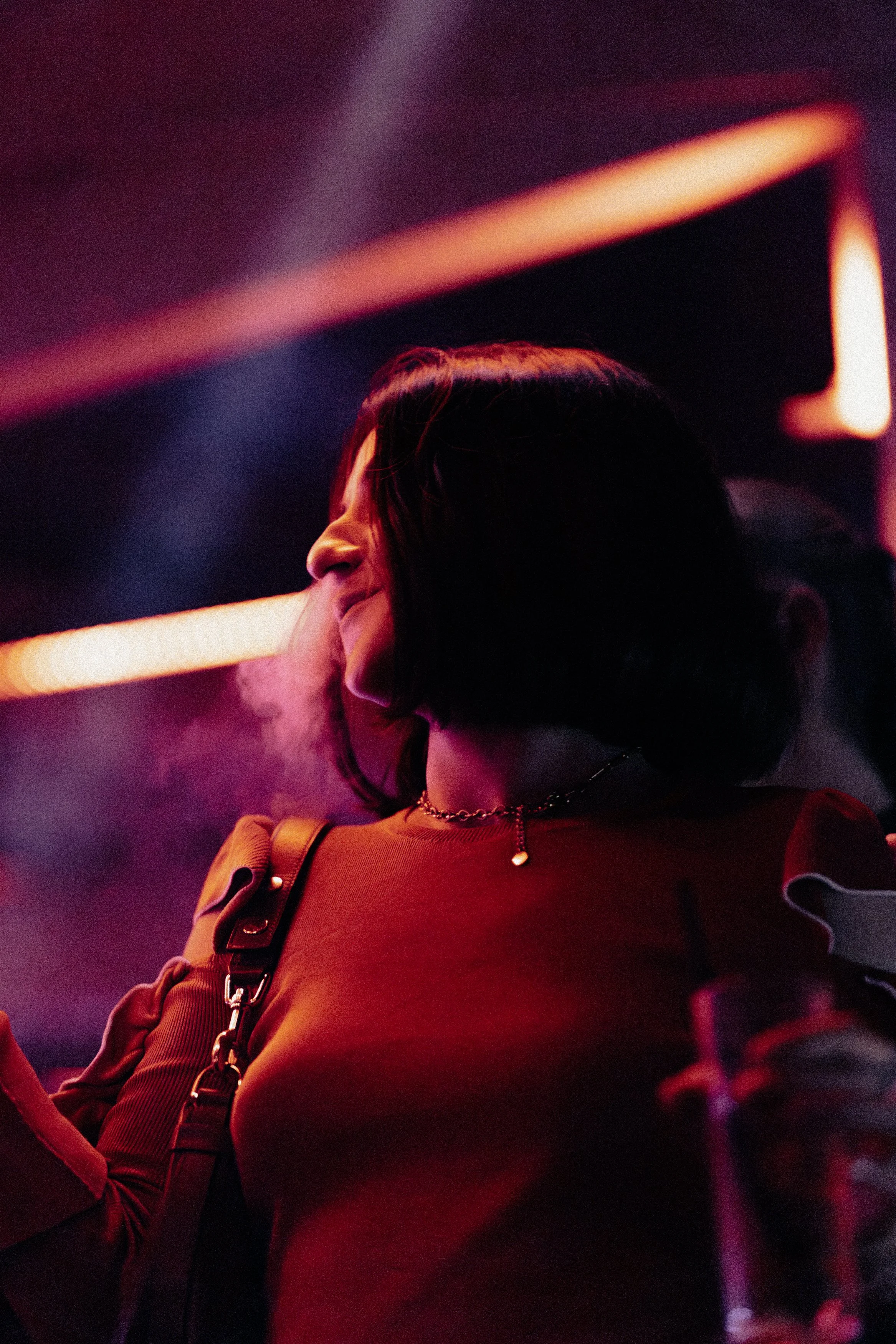 Une femme avec des cheveux courts, portant un haut rouge avec des épaulettes, un collier doré et un sac à bandoulière, dans un environnement sombre avec des lumières de néon rouges et violettes.