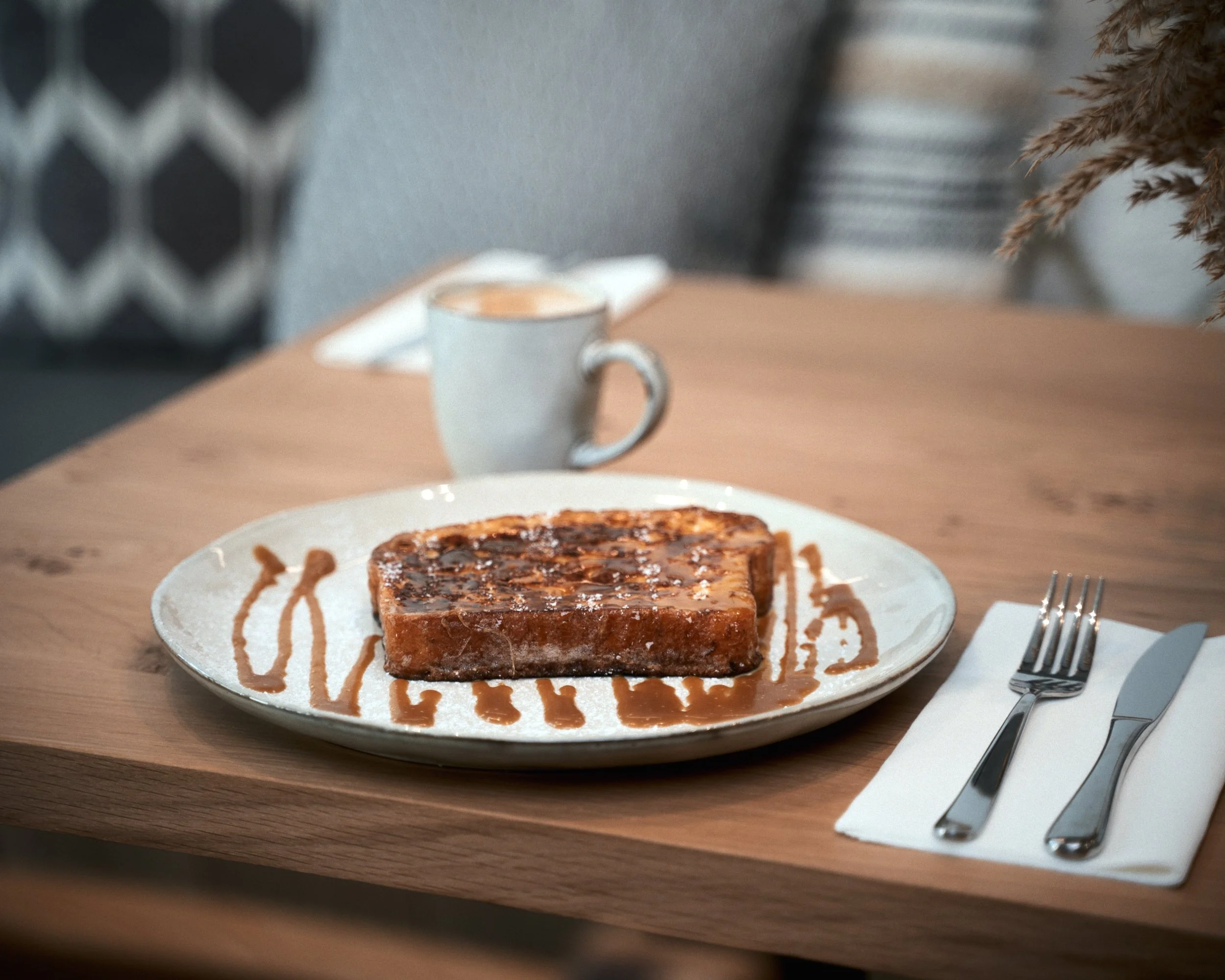 Tranche de pain perdu avec du sirop, une tasse à café en arrière-plan, une fourchette et un couteau sur une serviette blanche à droite, le tout sur une table en bois.