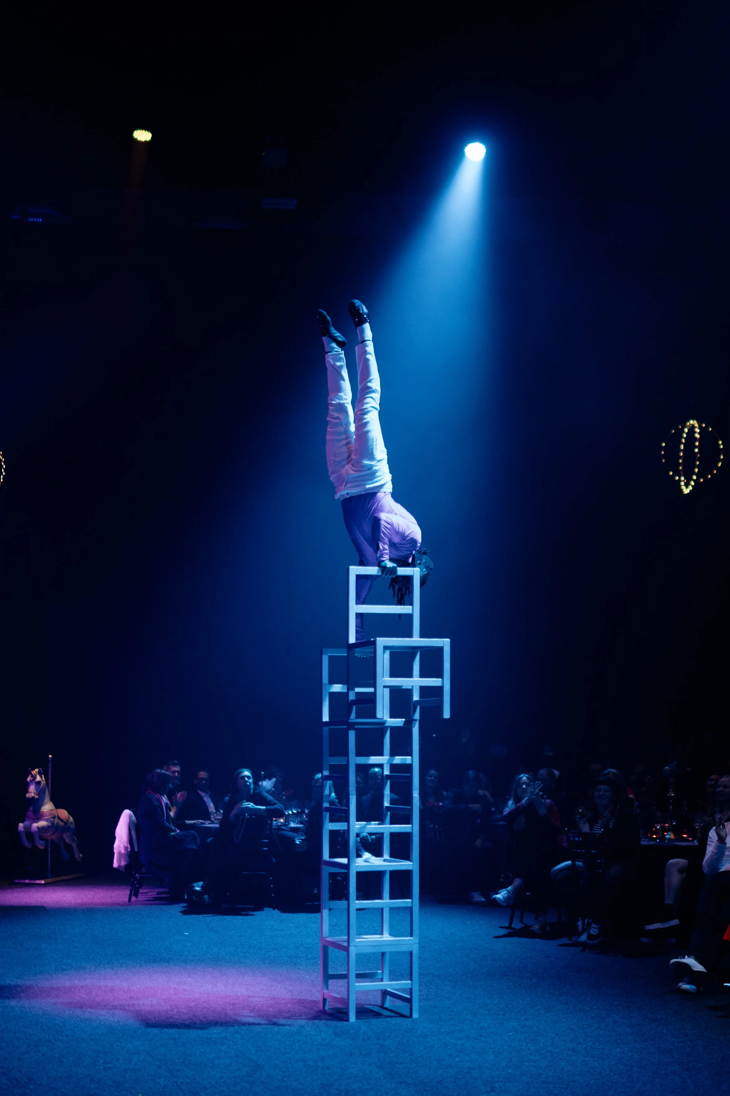 Un artiste de cirque en équilibre en position de tête sur une pile de chaises sous un éclairage bleu, avec un public assis en arrière-plan.