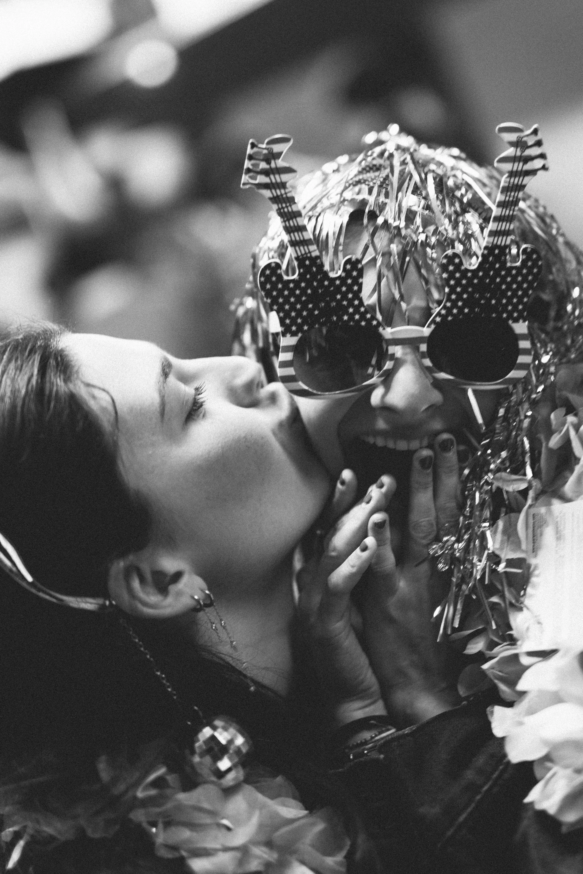 Une femme embrasse un homme portant des accessoires de fête, notamment des lunettes et un gâteau décoratif, lors d'une célébration.