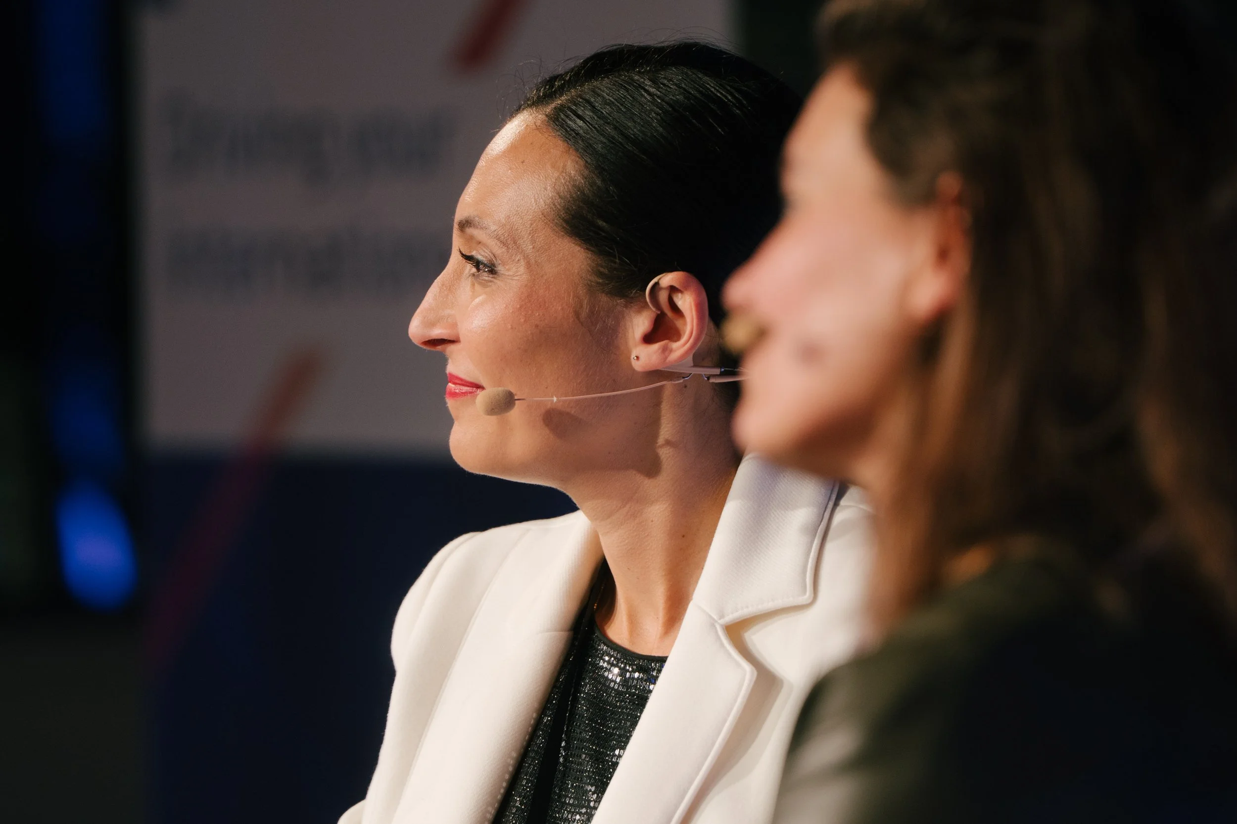 Deux femmes en train de parler lors d'une conférence, l'une portant un micro-cravate beige, l'autre en arrière-plan, floue.