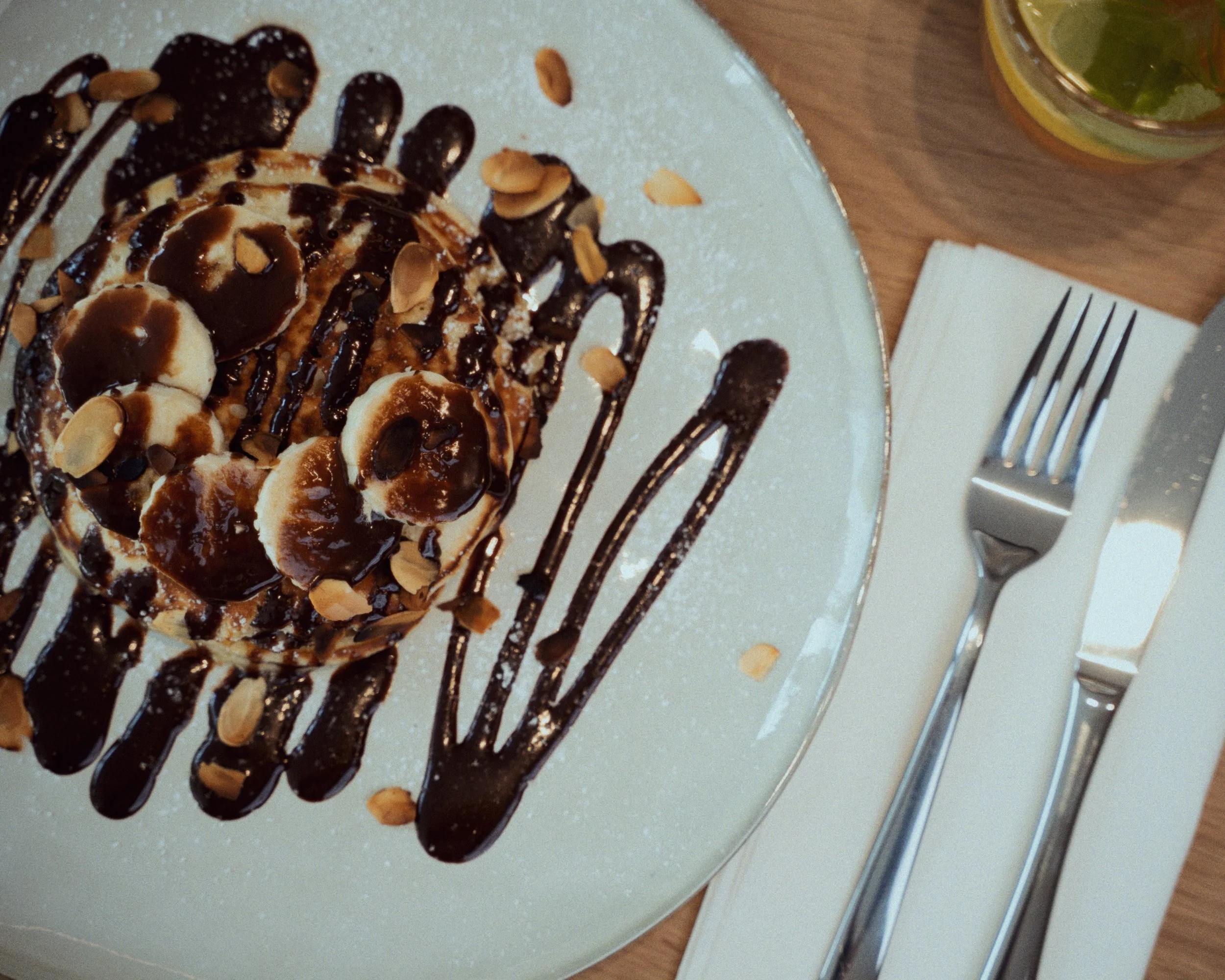 Gâteau aux bananes, garniture de chocolat fondu, amandes effilées, et sauce au chocolat, servi sur une assiette blanche avec des couverts à côté.