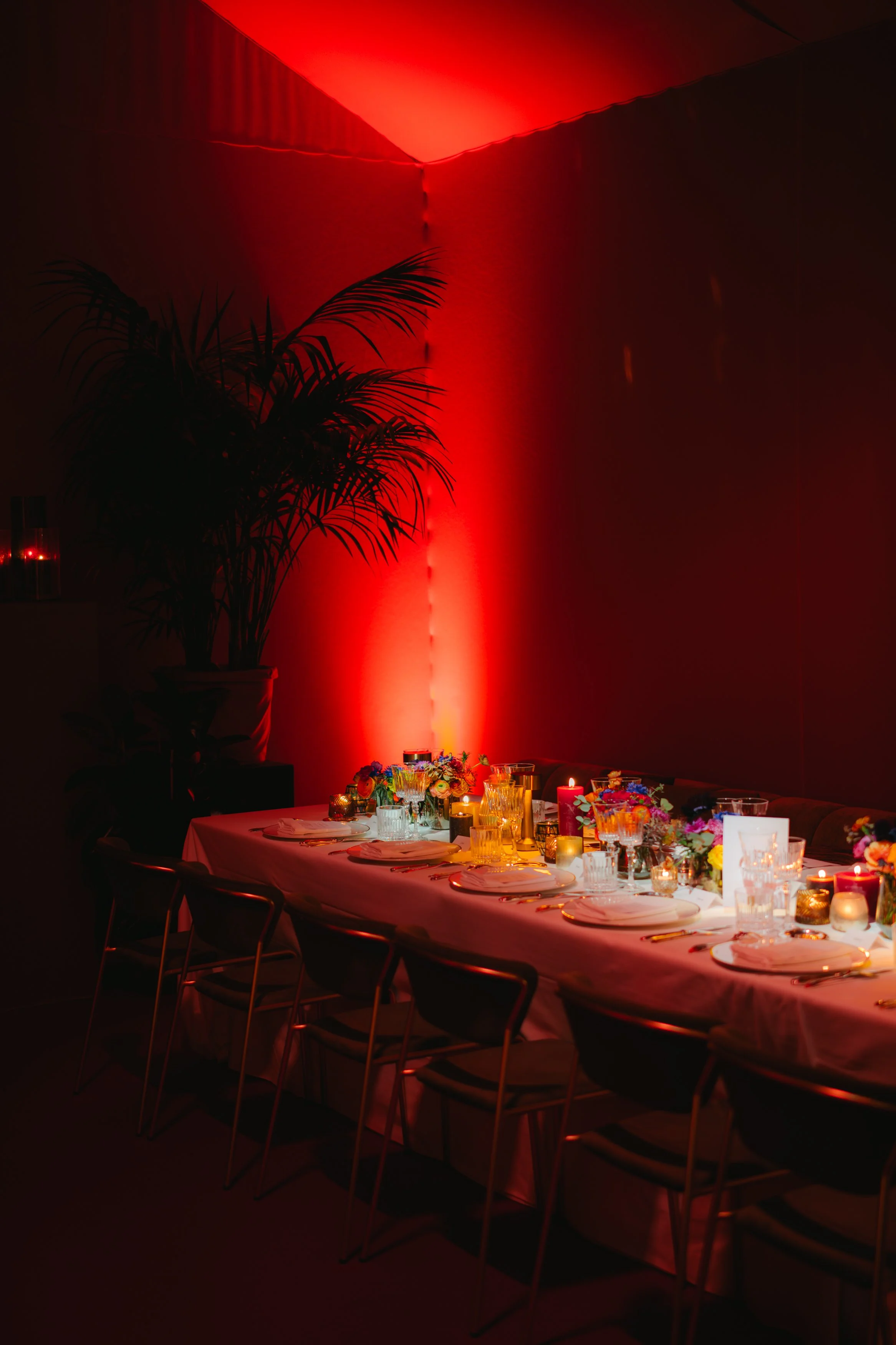 Table de fête décorée avec des fleurs, bougies et vaisselle dans une pièce éclairée en rouge, avec une grande plante verte en arrière-plan.