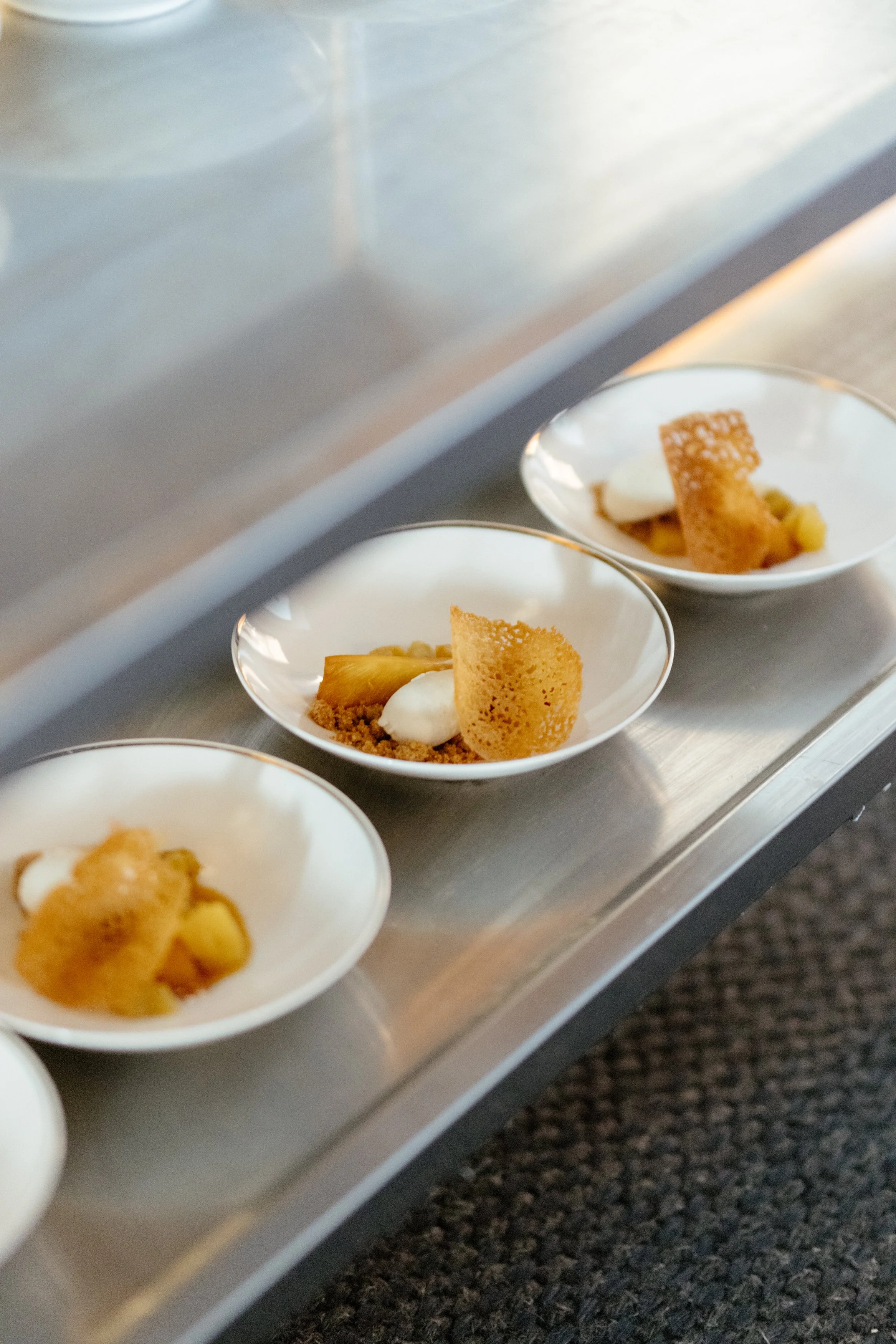 Desserts en portions individuelles sur une plaque en acier inoxydable, comprenant une pâte cuite, une cuillerée de crème ou de mousse, et une fine biscuit croustillant.