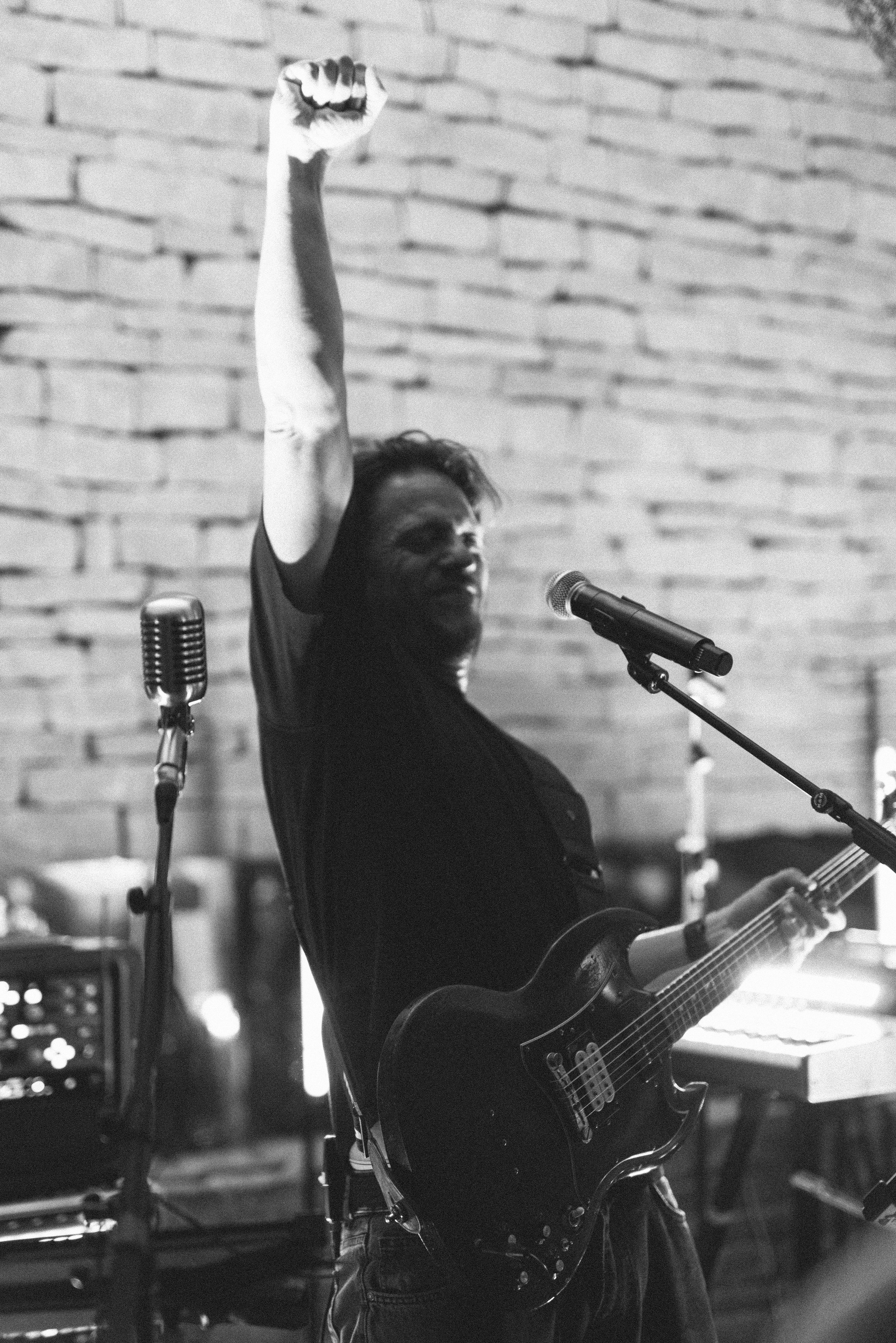 Un musicien avec une guitare électrique, levant le bras en l'air, enregistrement dans un espace intérieur avec un mur en briques.