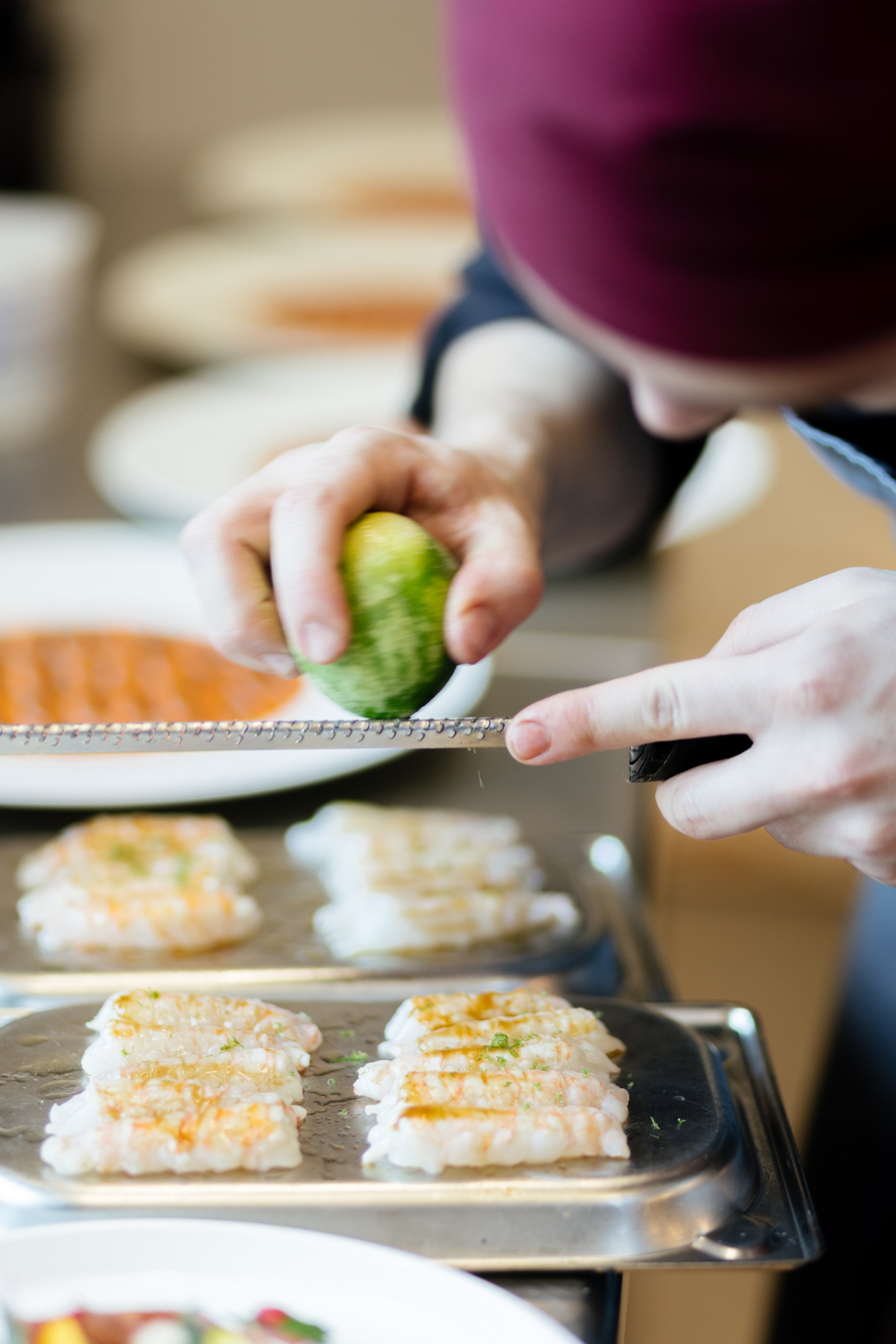 Une personne qui prépare des sushis, en train de râper un concombre pour garnir des roules de sushi sur une plaque en acier inoxydable.
