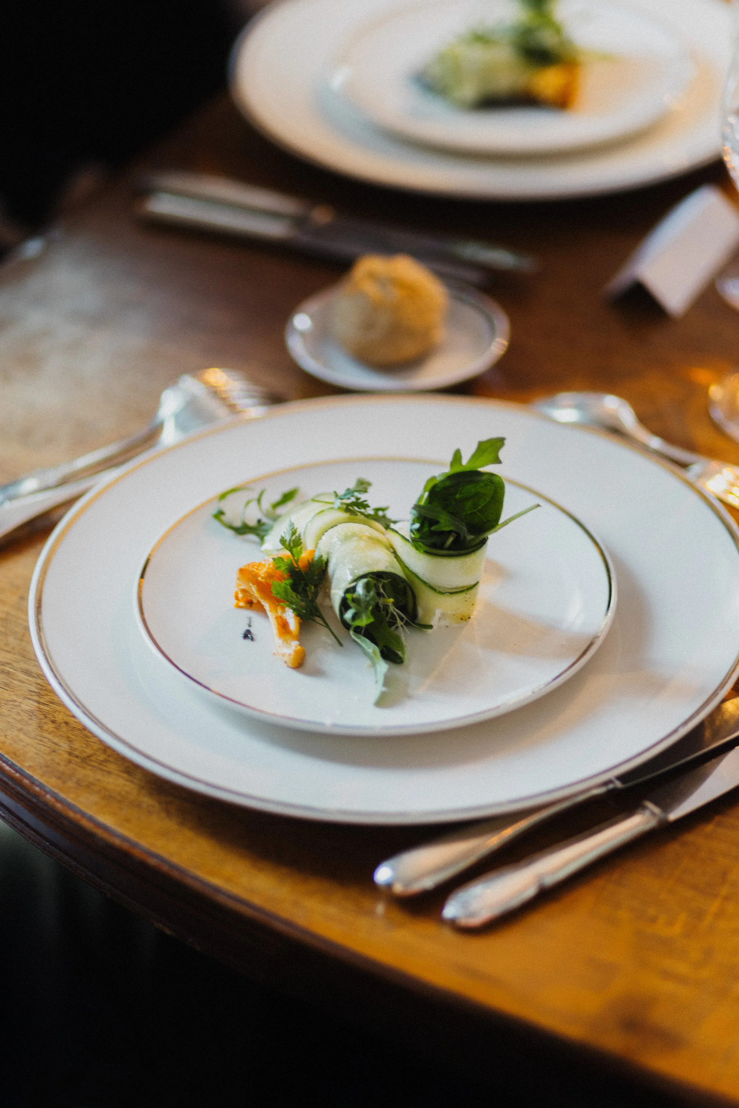 Assiette fine avec rouleaux de concombre et légumes verts, garnie de feuilles de salade, sur une table en bois avec éléments de vaisselle en argent.