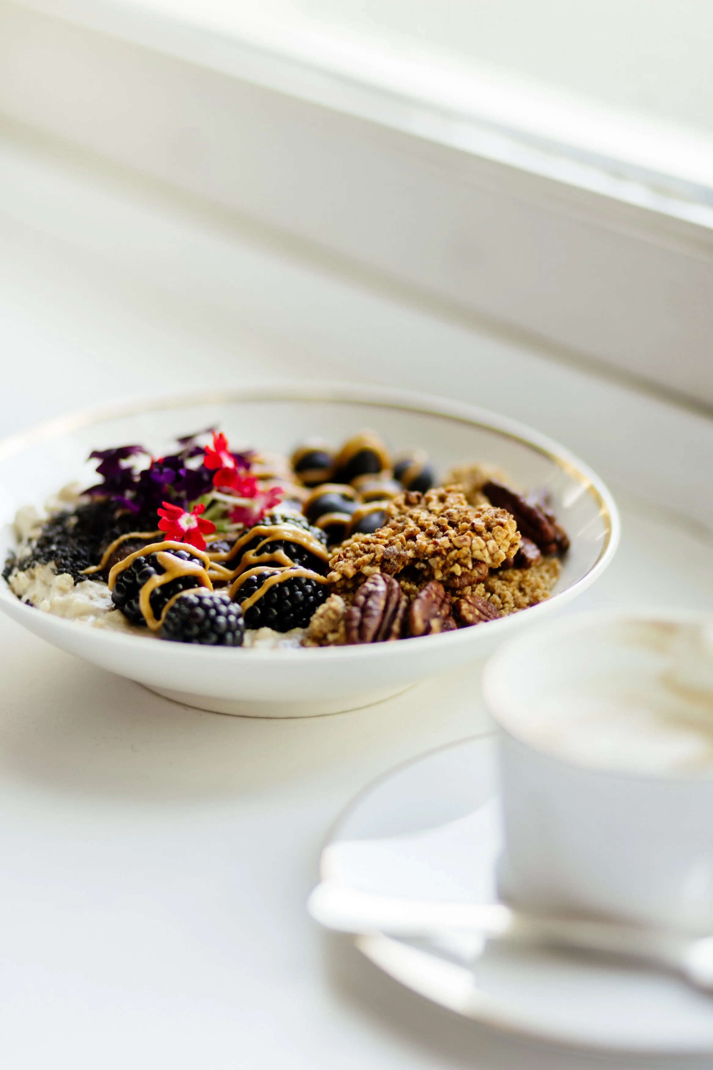 Un bol de céréales avec des mûres, des fruits secs et du miel, à proximité d'une tasse de lait ou de crème, sur une surface blanche près d'une fenêtre.