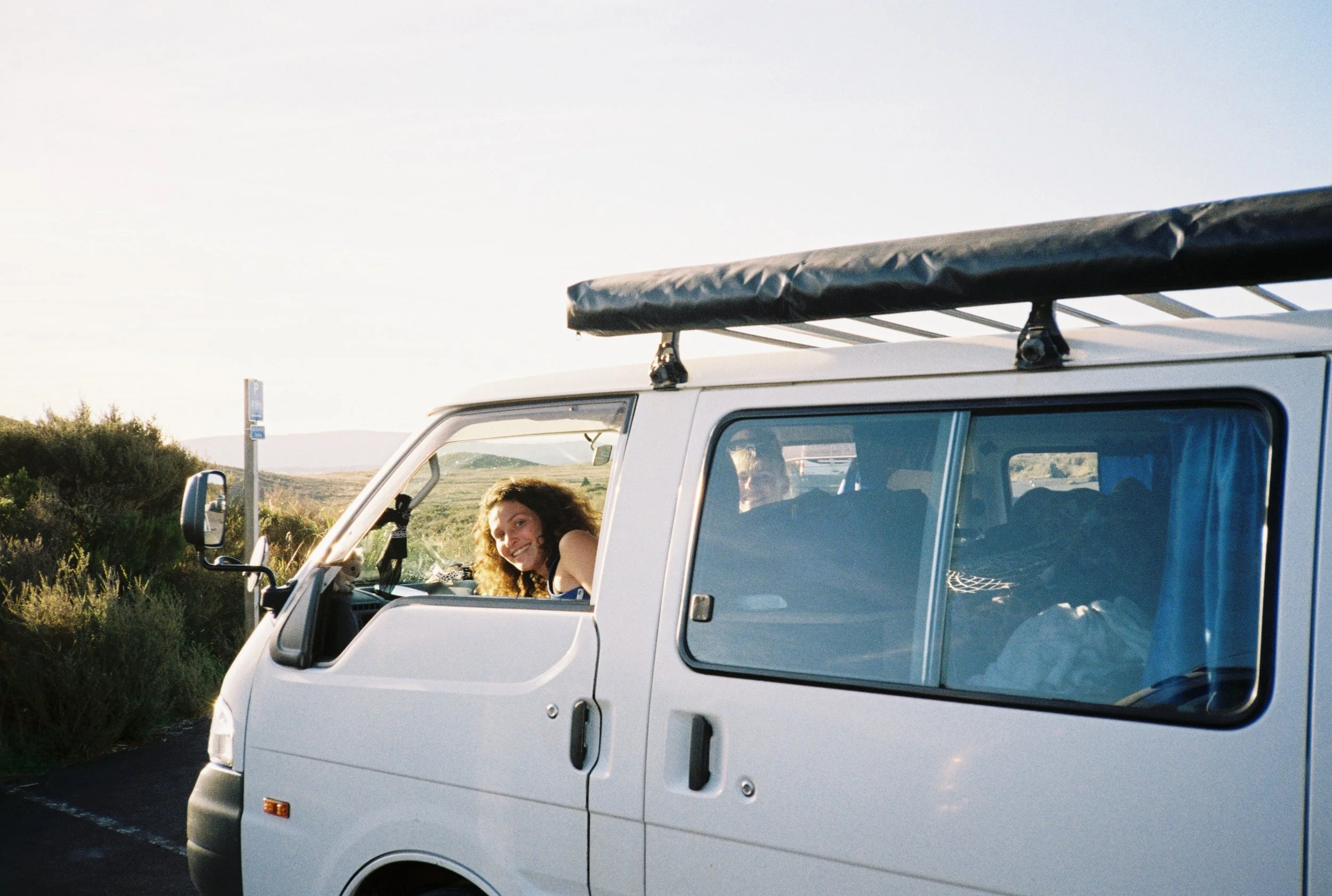 Une femme souriante qui regarde par la fenêtre d'une camionnette blanche en plein air, avec un paysage rural en arrière-plan et un ciel clair.