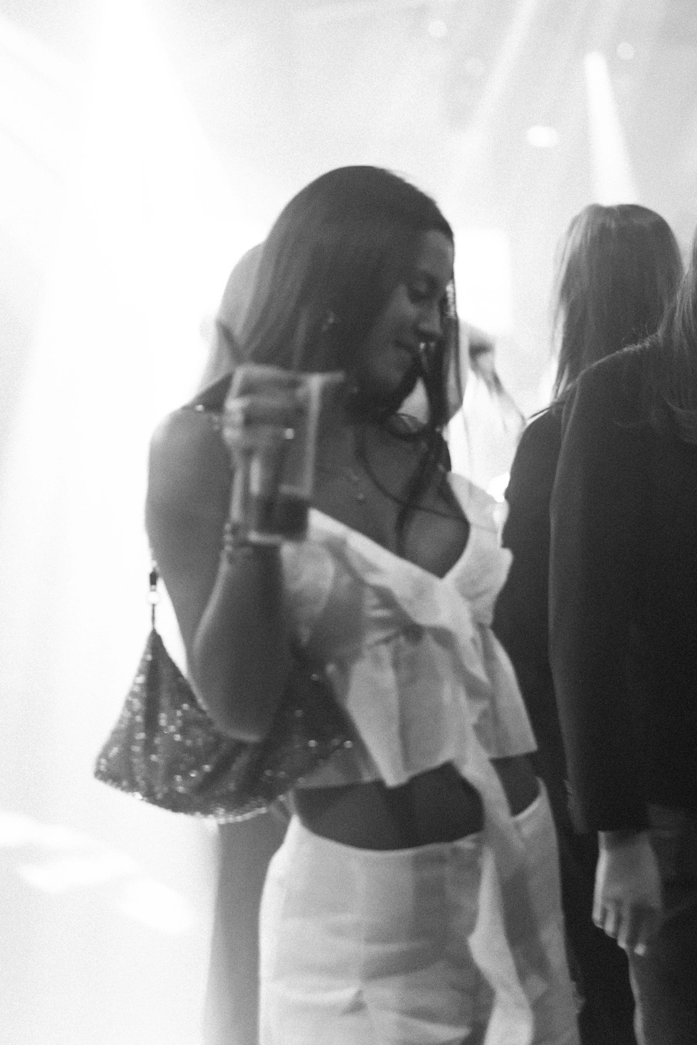 Une femme dans un décor sombre, portant une robe légère, un sac à main, une veste et un vêtement noir visible autour de la taille, dans une ambiance de fête ou de club.