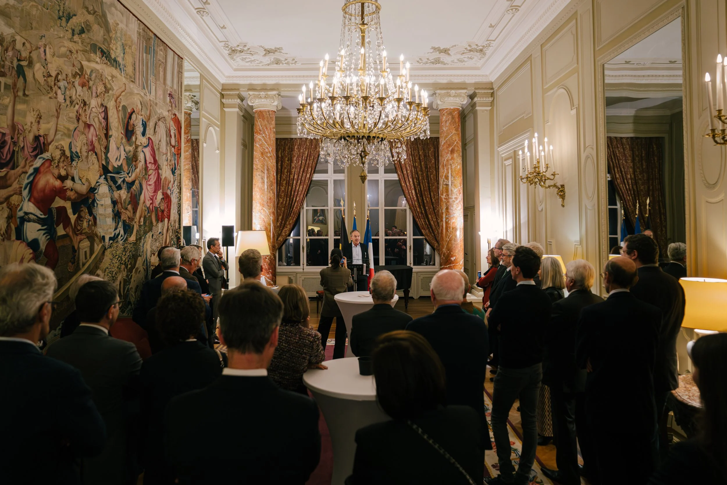 Une réunion ou un discours formel dans une salle élégante avec un chandelier chandelier au plafond, des murs ornés de tapisseries et des grandes fenêtres avec des rideaux. Plusieurs personnes écoutent un homme au centre qui parle derrière un pupitre.