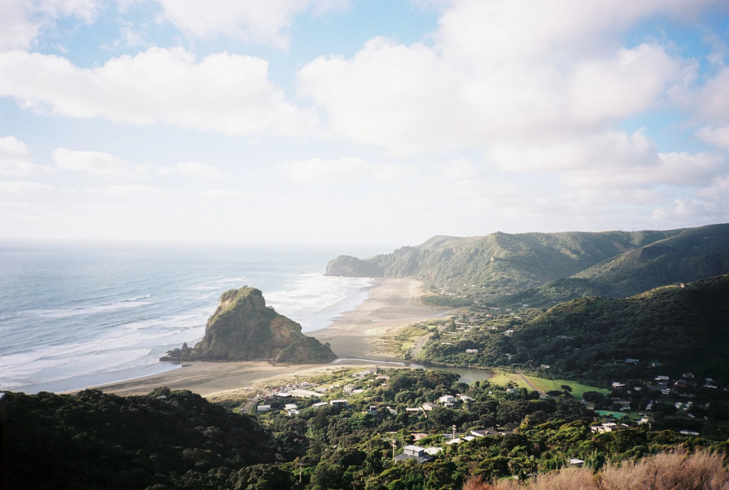 Paysage côtier avec plages, montagnes verdoyantes, petit village et océan sous ciel partiellement nuageux