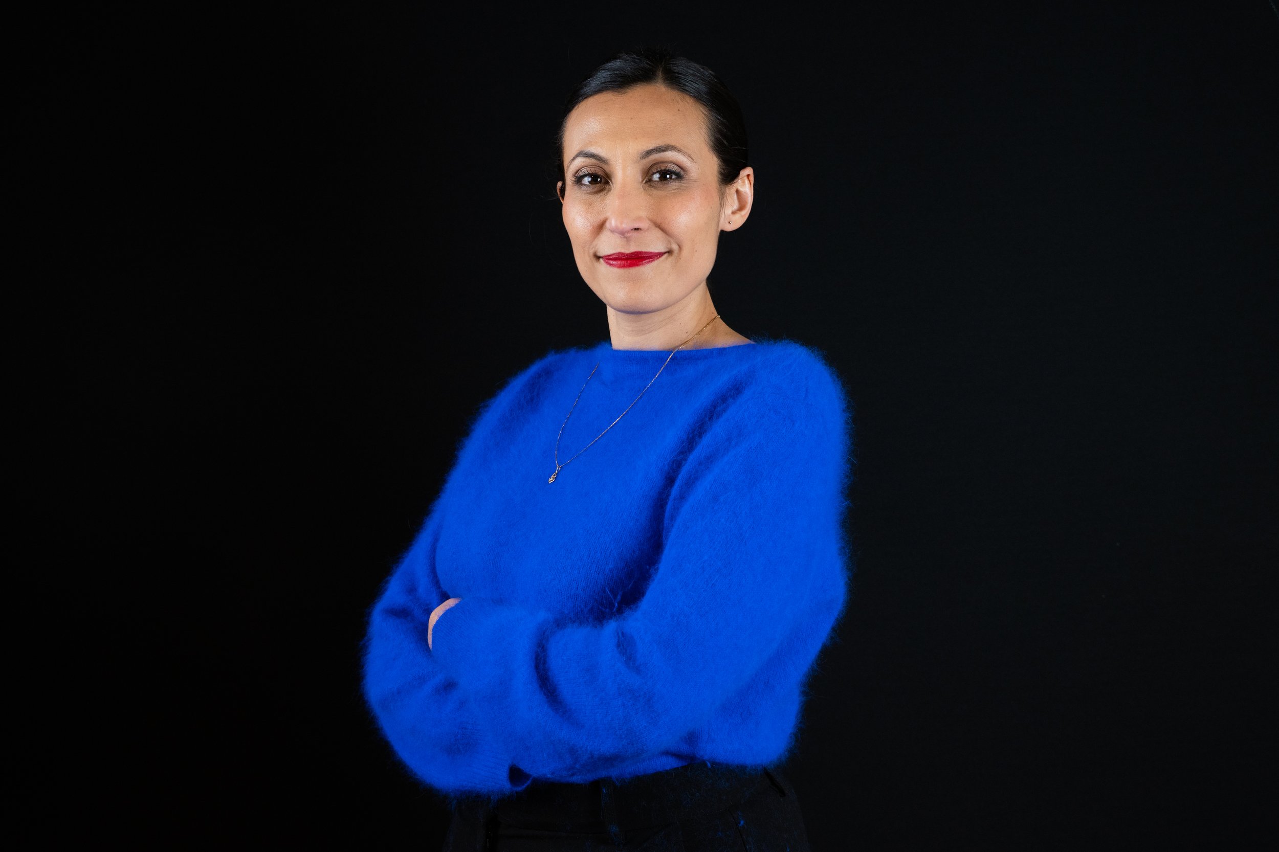 Femme souriante portant un pull bleu vif, bras croisés, sur fond noir.