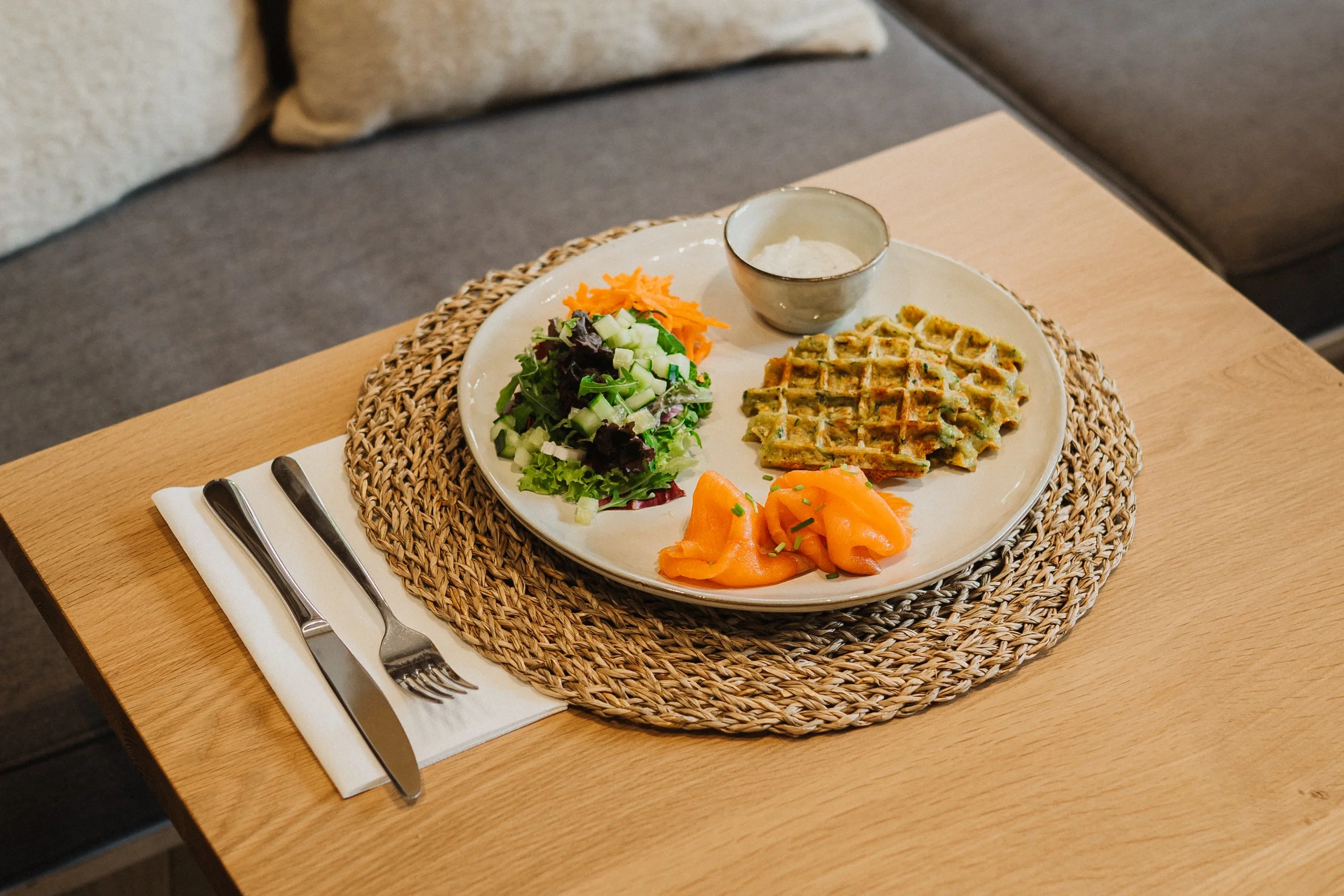 Assiette de nourriture avec une salade verte, des soupes de saumon fumé, des gaufres aux herbes, et une sauce dans un bol, sur une table en bois avec une nappe en osier.