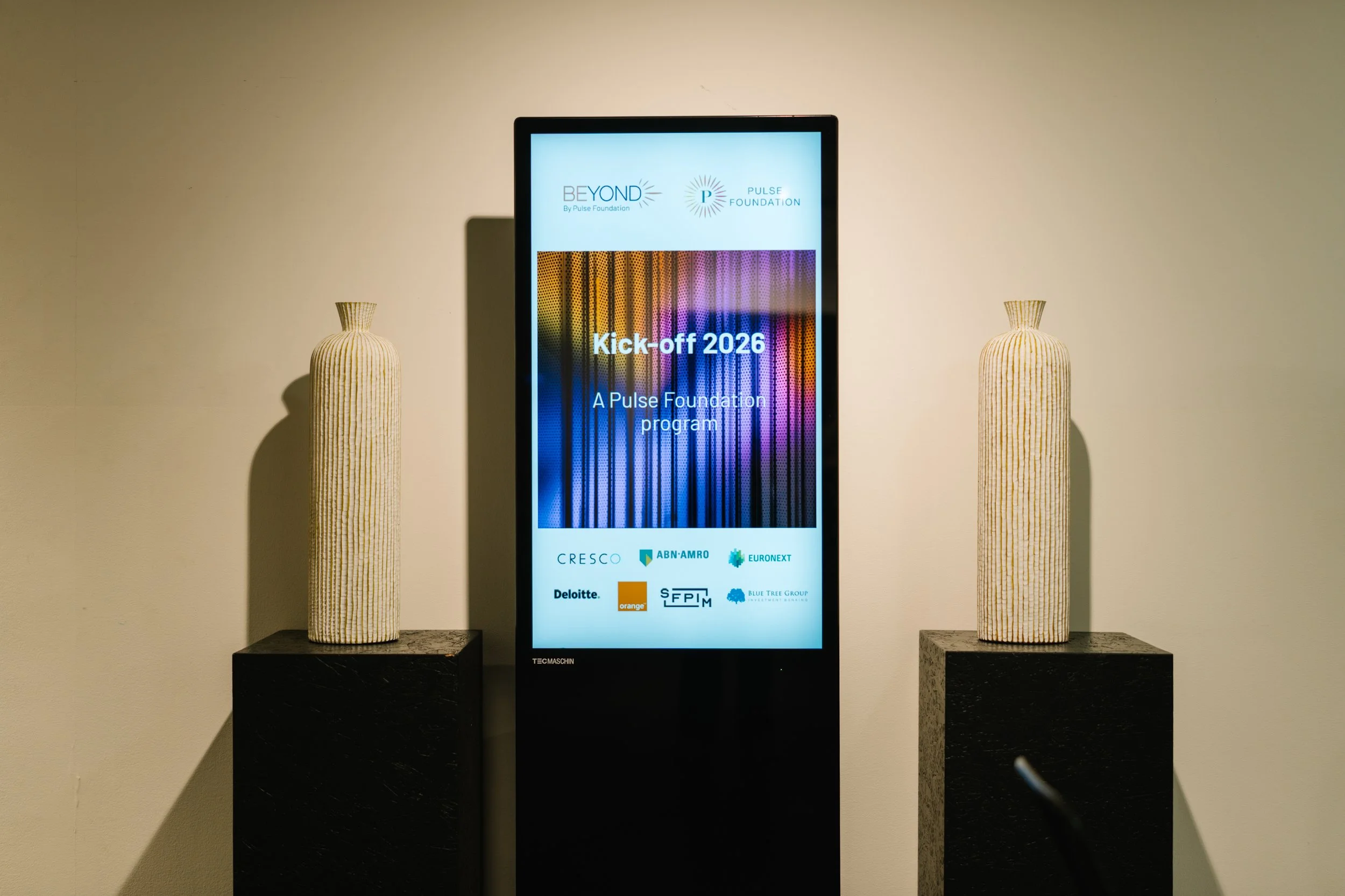 Un écran digital affichant l'annonce du lancement de 2026 pour le programme Pulse Foundation, avec deux vases décoratifs jaune clair placés sur des socles noirs de chaque côté.