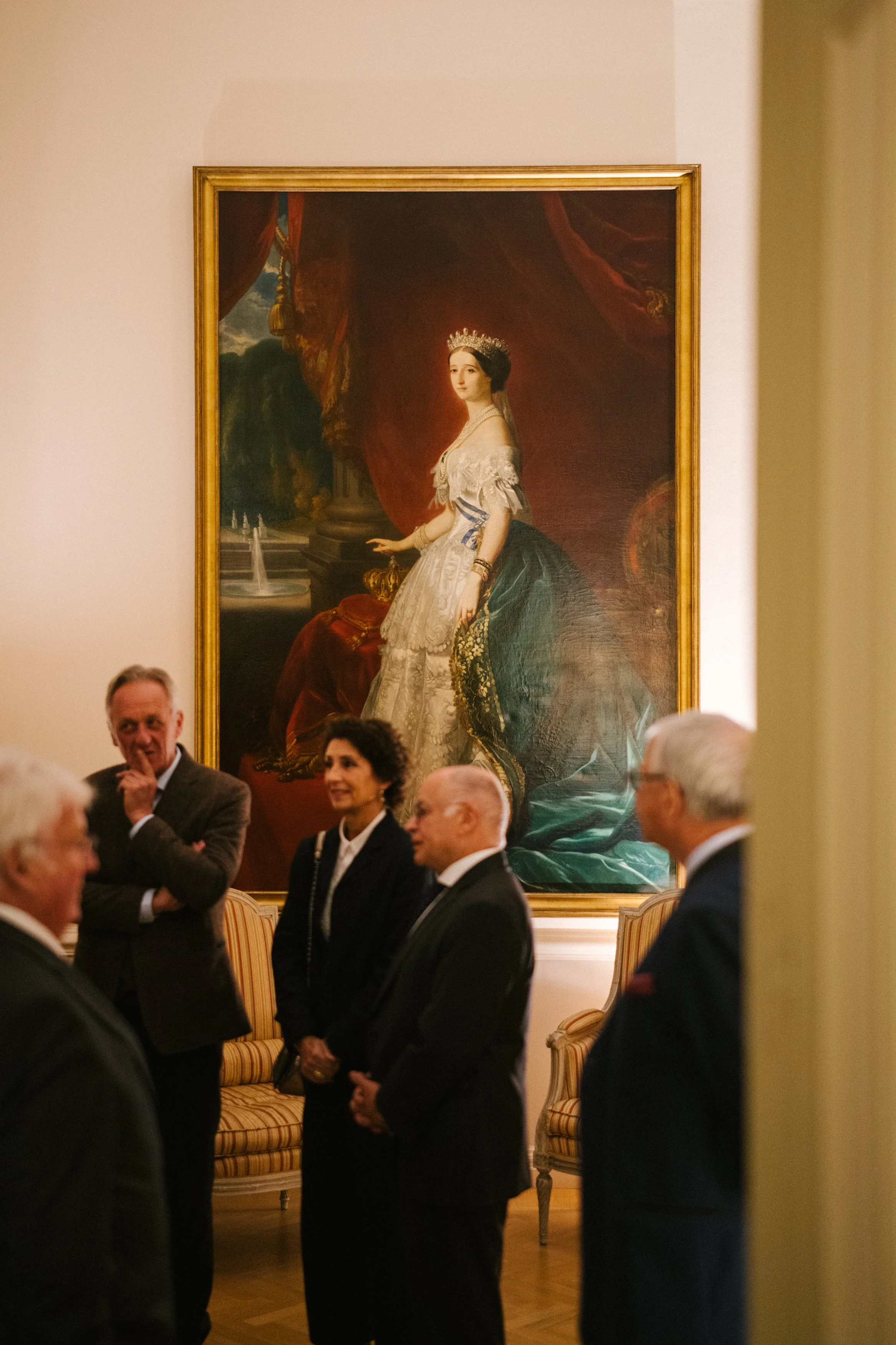 Une salle d'art avec un grand tableau de portrait d'une femme en robes anciennes, entourée de plusieurs personnes en conversation.
