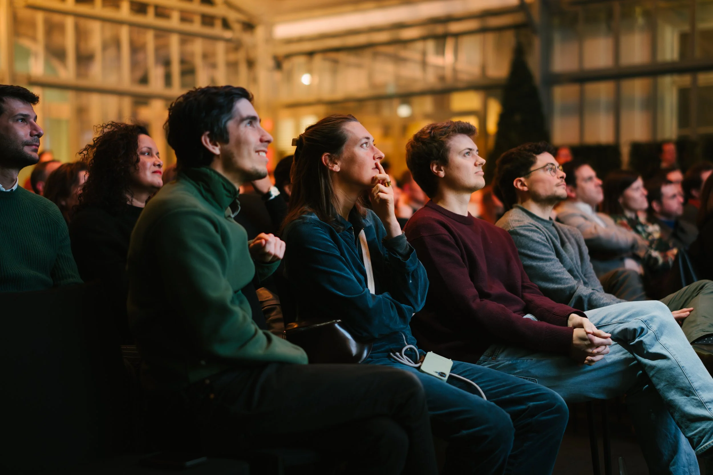 Groupe de personnes assises, regardant un spectacle ou une conférence, dans une salle avec une ambiance chaleureuse.